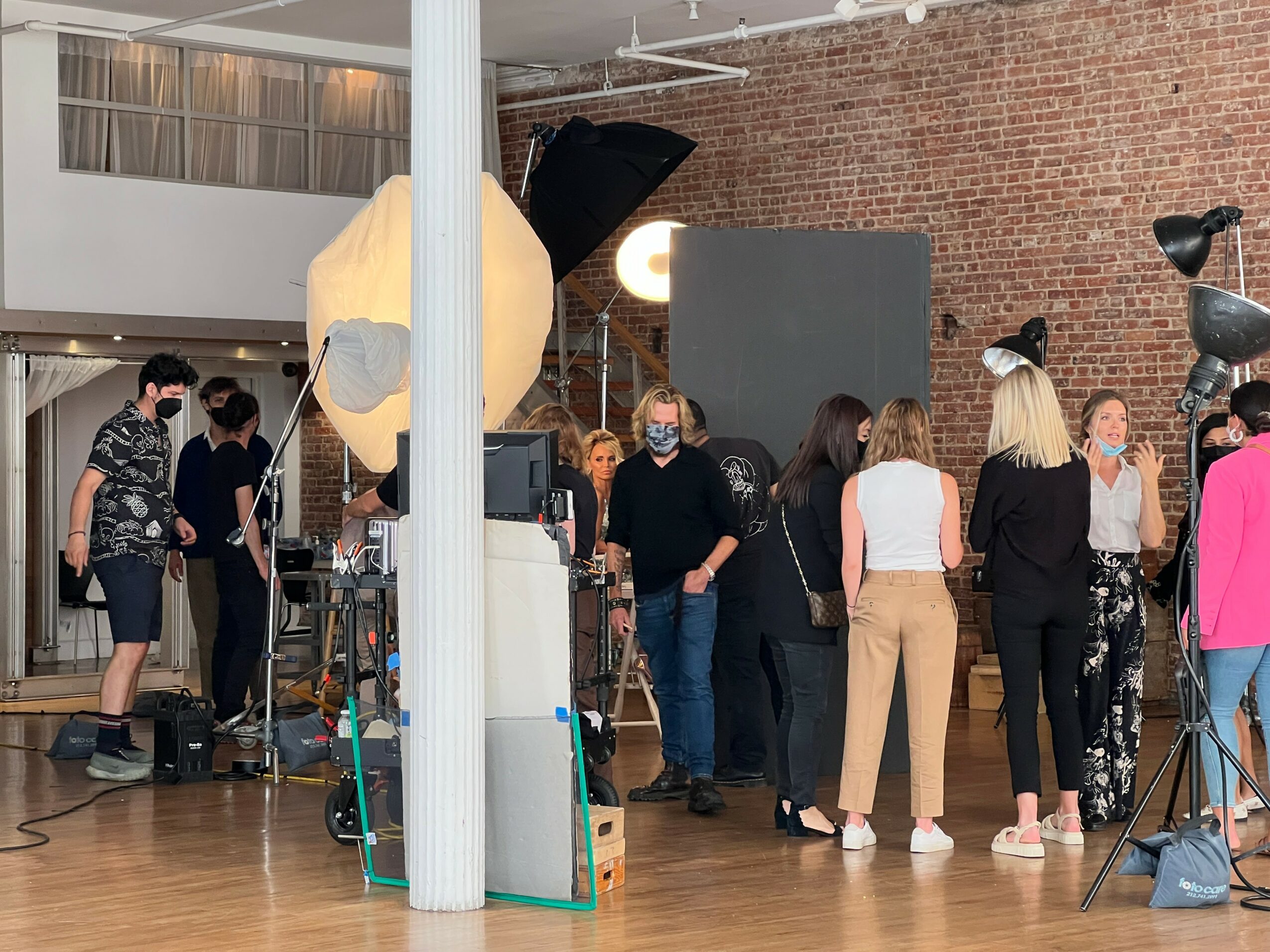 Group of people in a studio with brick wall, equipment, and a large sculpture, some wearing masks, engaged in conversation.
