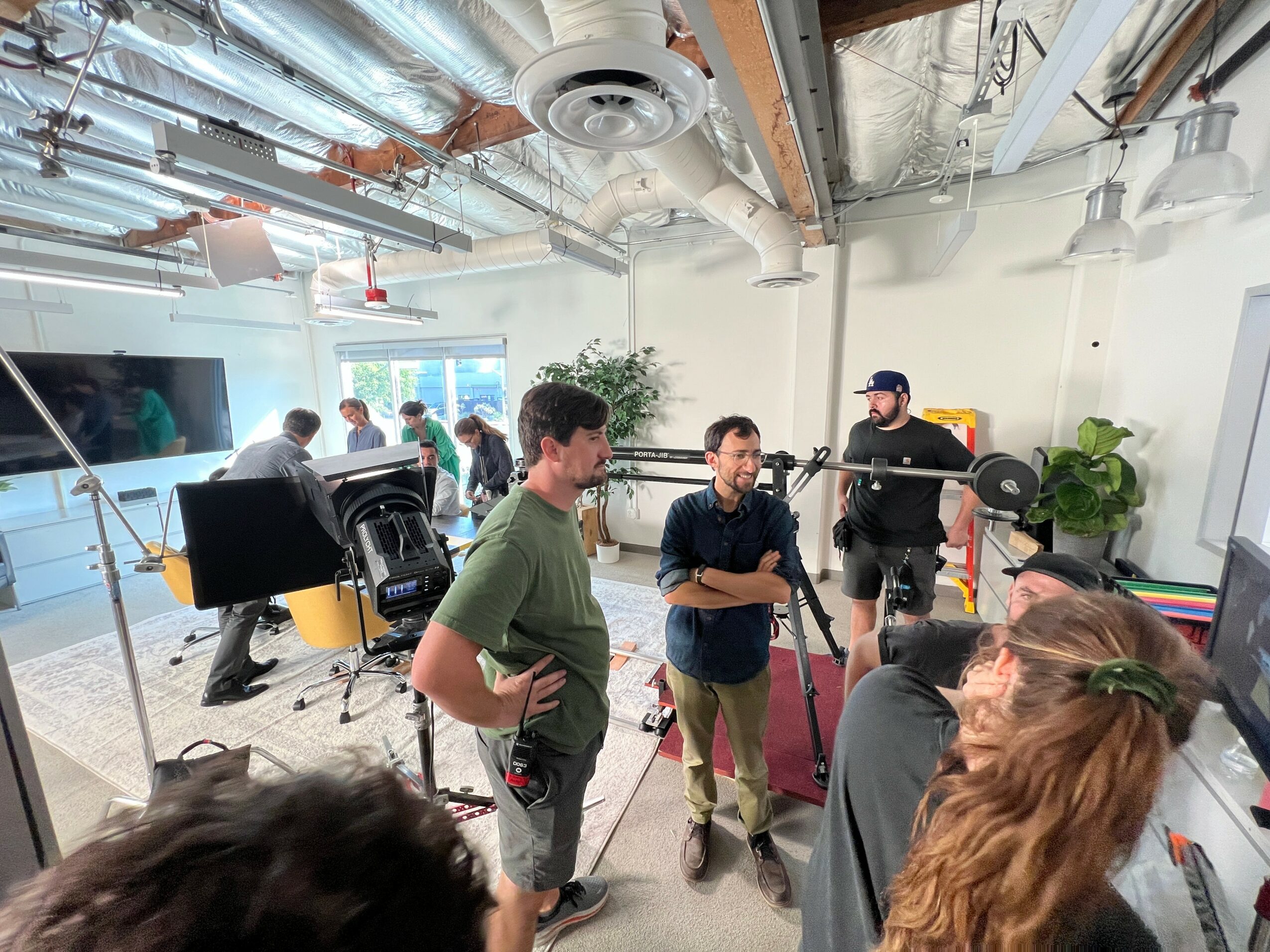 Group of people in a modern, open office space with exposed ceiling pipes, large windows, and plants, engaged in conversation.