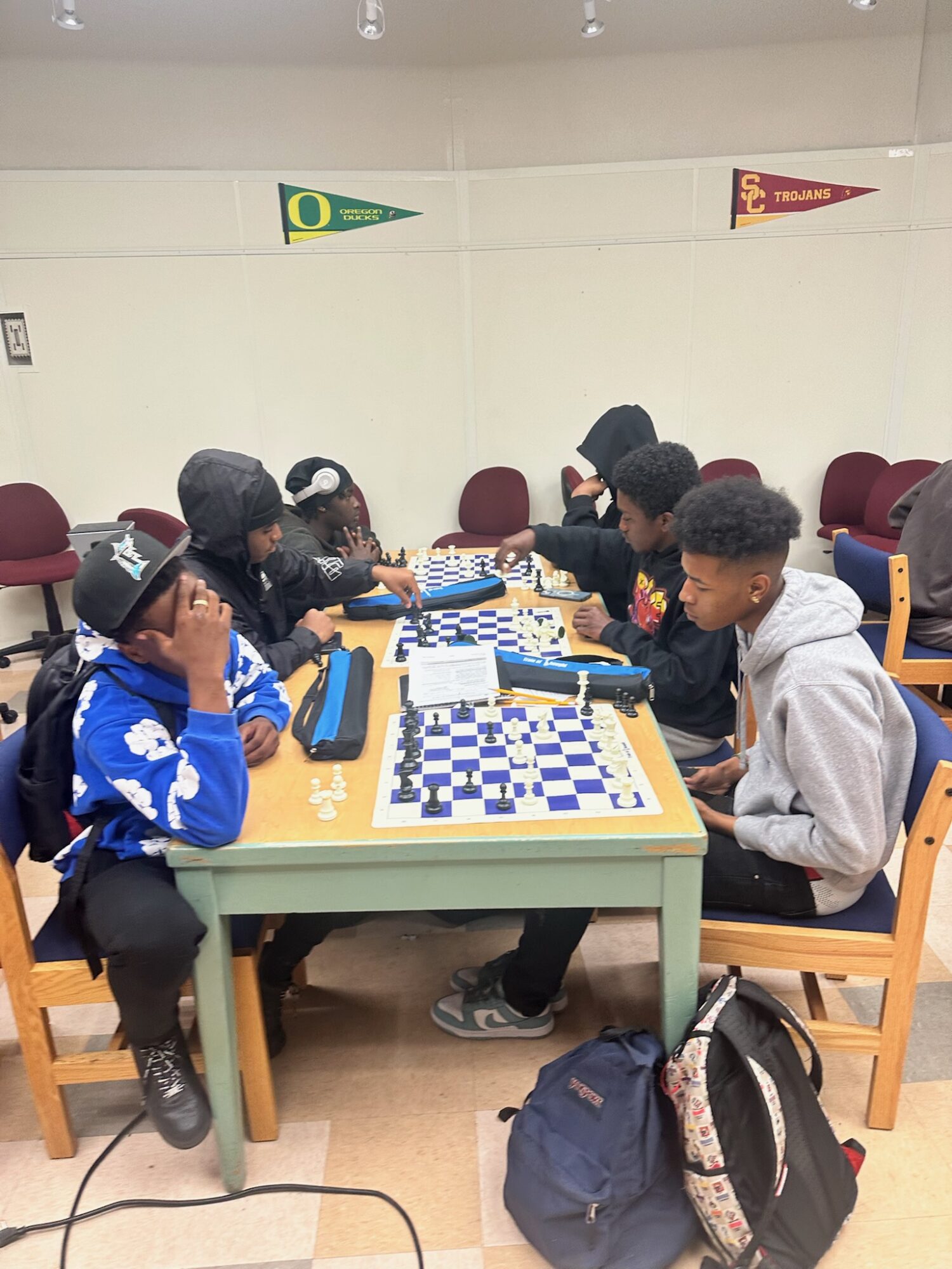 Two groups of children playing chess at tables in a room with flags on the wall, backpacks on the floor, and chairs around the tables.