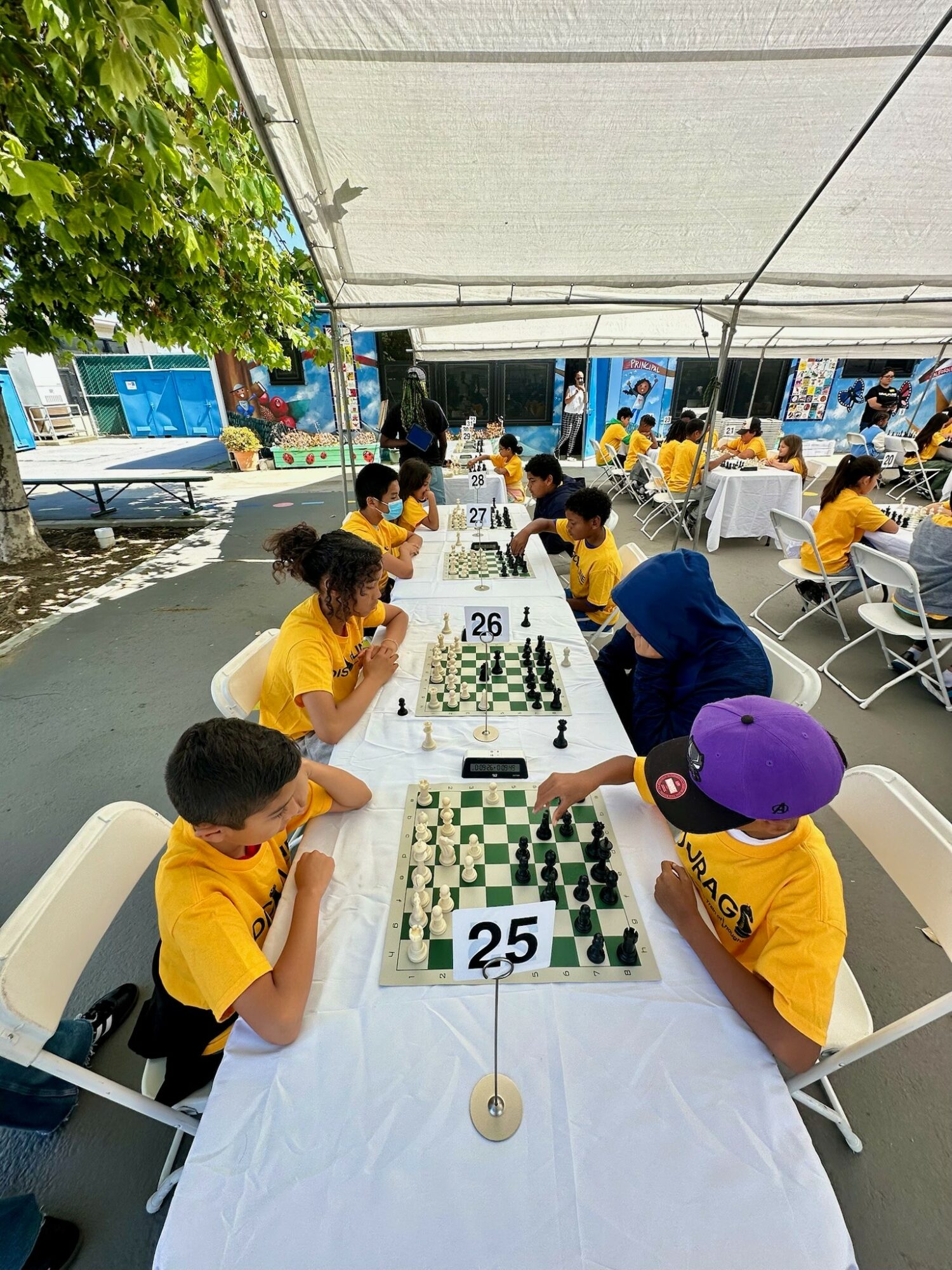 Children in yellow shirts playing chess at a long table outdoors under a canopy, with trees and people in the background.