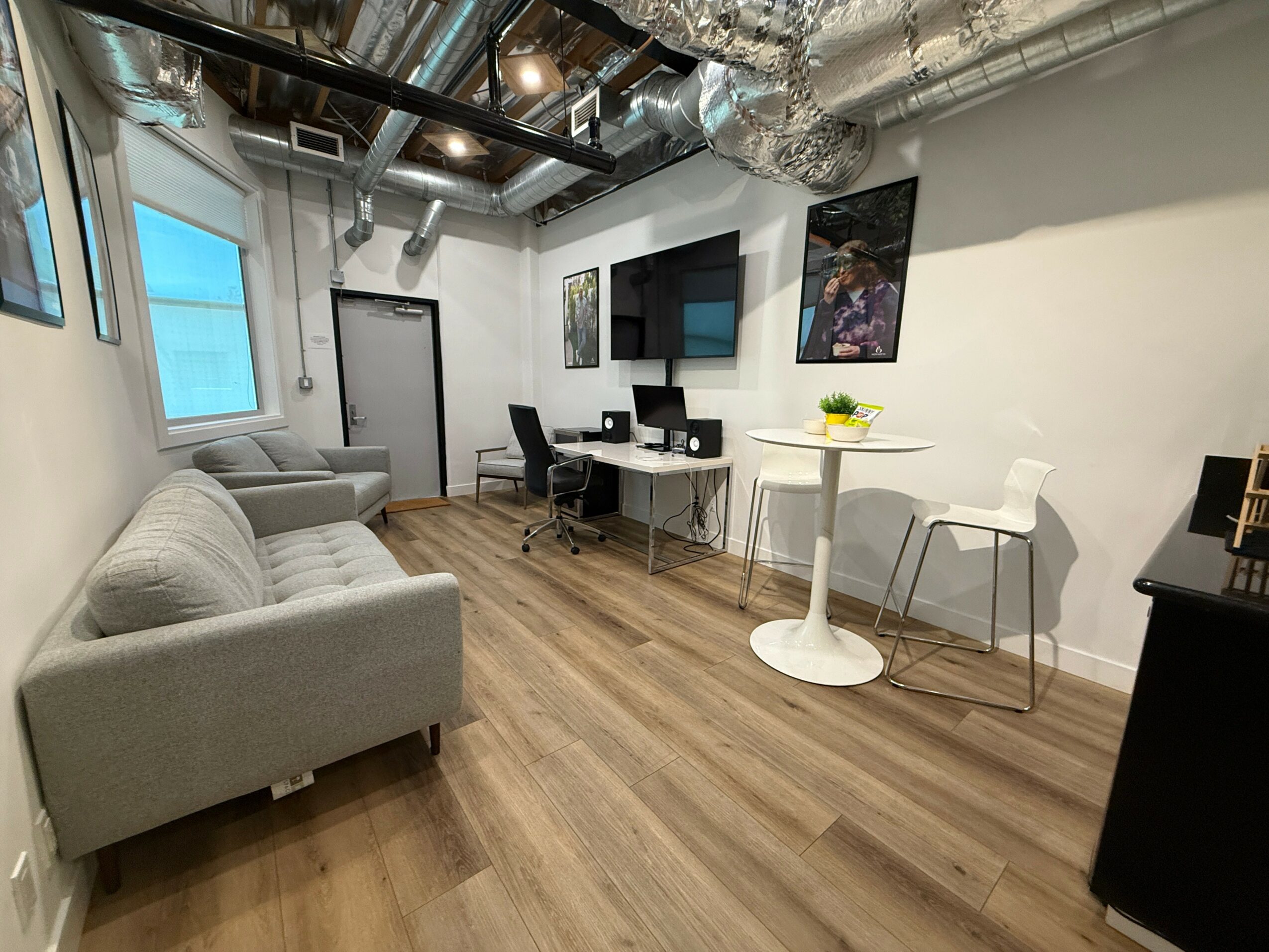 Office with a gray sofa, desk with computer, wall-mounted TV, and a small round table with two chairs, wooden floor.