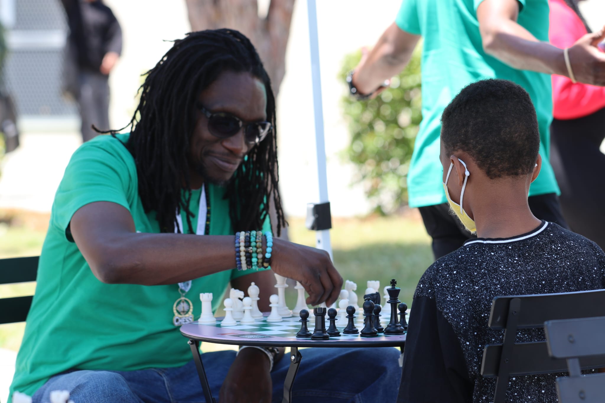Woman with dreadlocks and sunglasses playing chess outdoors with a young boy wearing a face mask, other people in background.