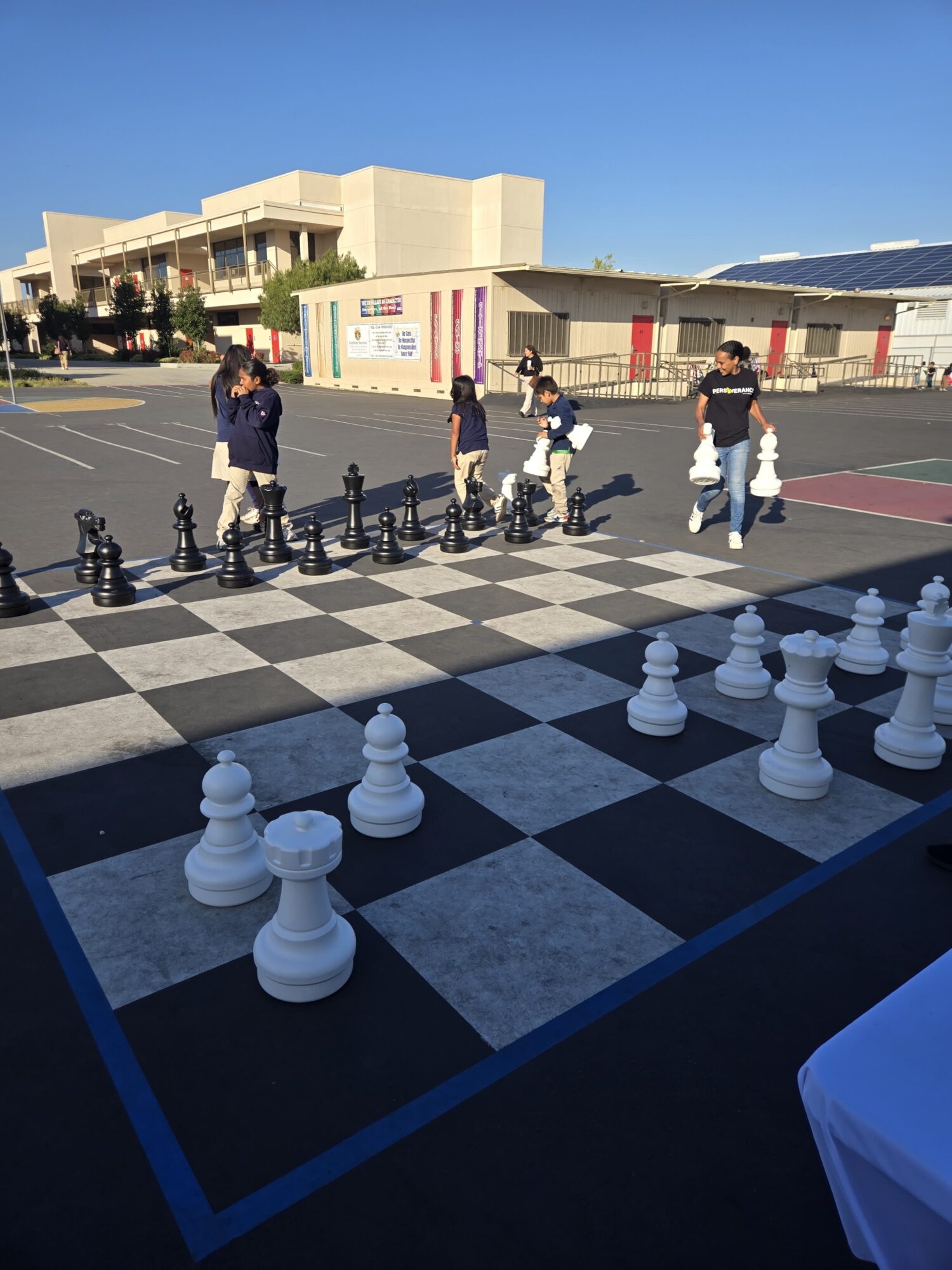 Group of people walking across a large outdoor chessboard with oversized chess pieces, in a sunny open area.