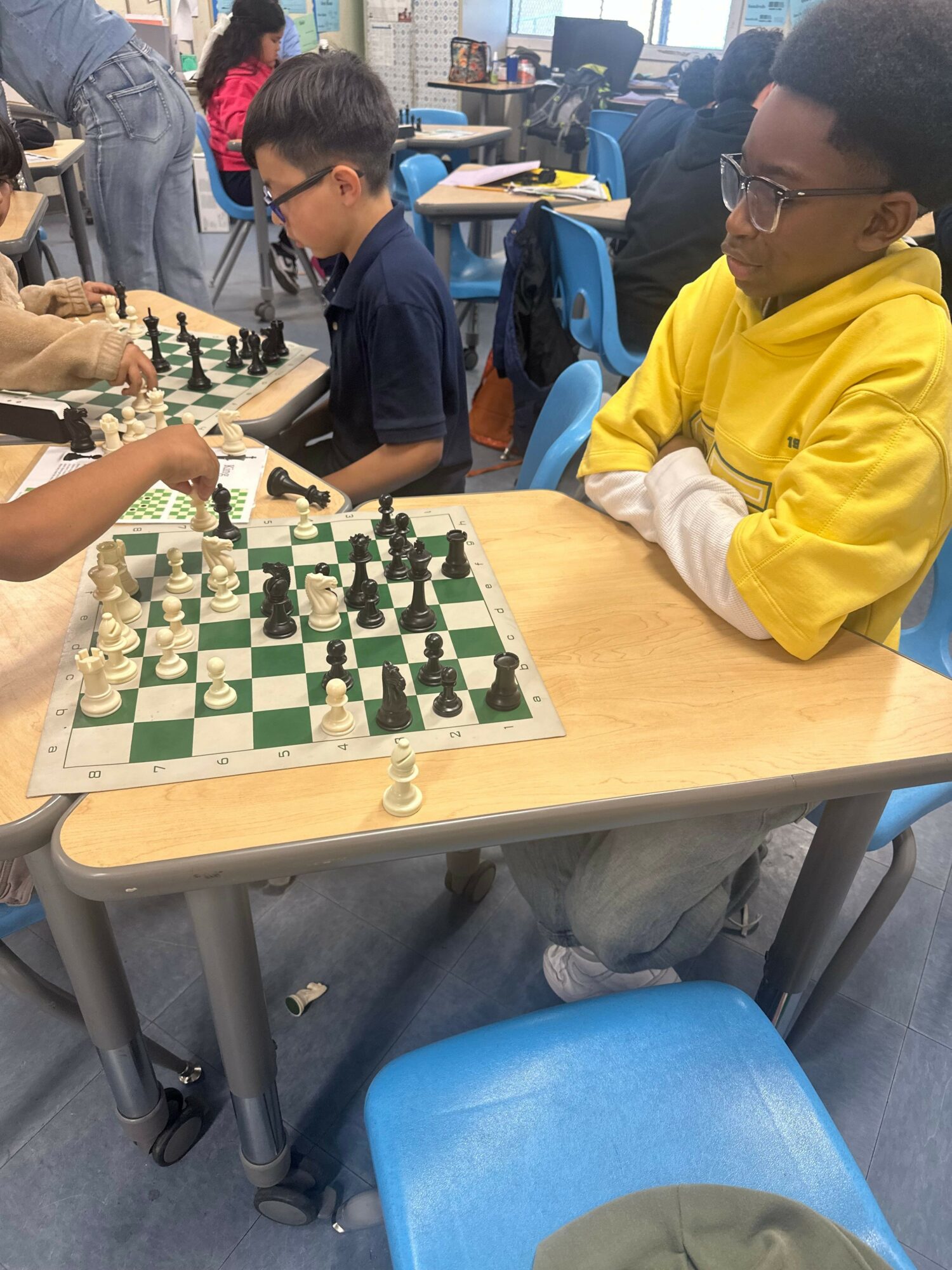Two children playing chess at a table in a classroom, with a teacher observing nearby, blue chairs around the table.