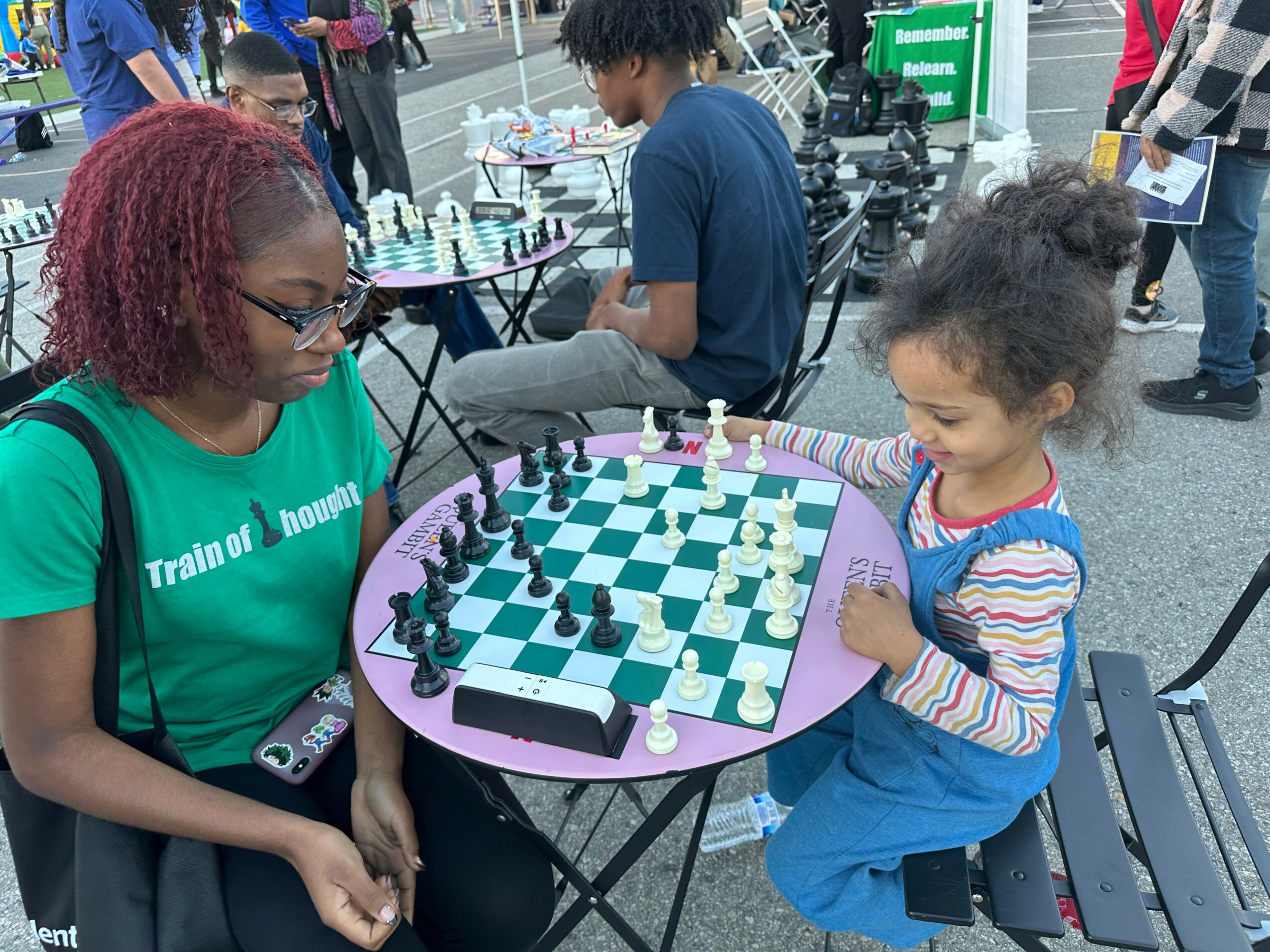 Two girls playing chess outdoors at a table, with other people and tables in the background.