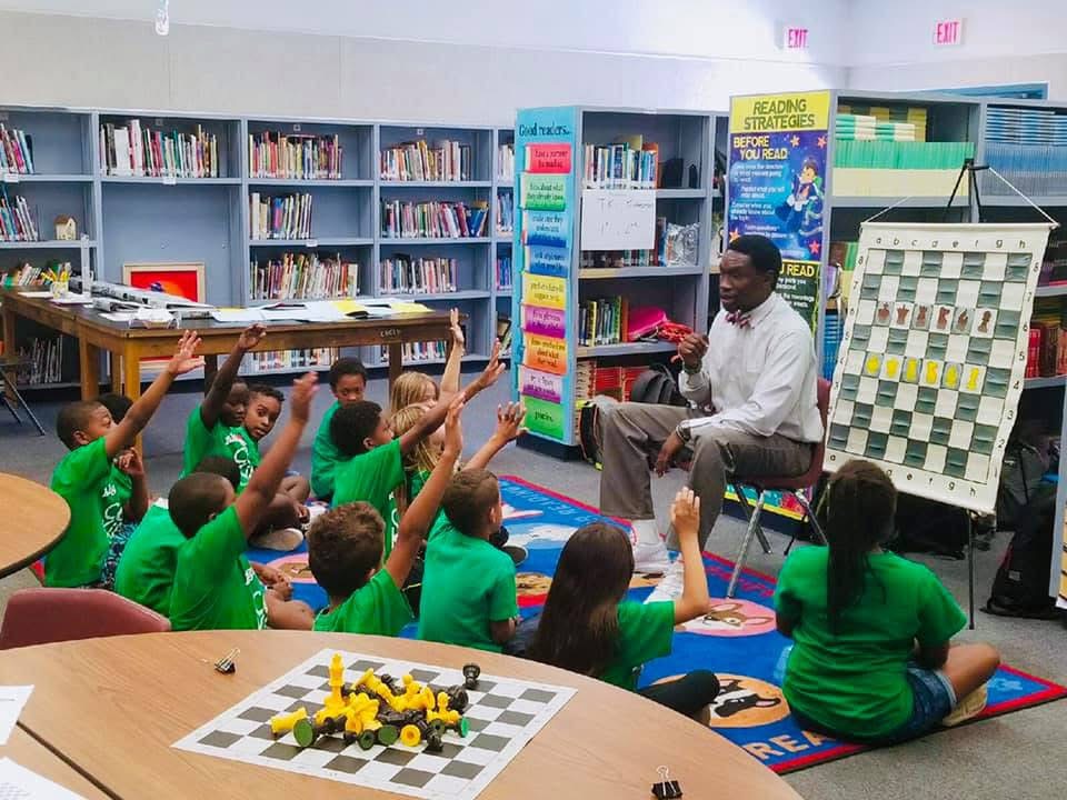 Children in green shirts sit on a colorful carpet in a library, raising hands while a man reads aloud. Bookshelves and a chessboard are visible.