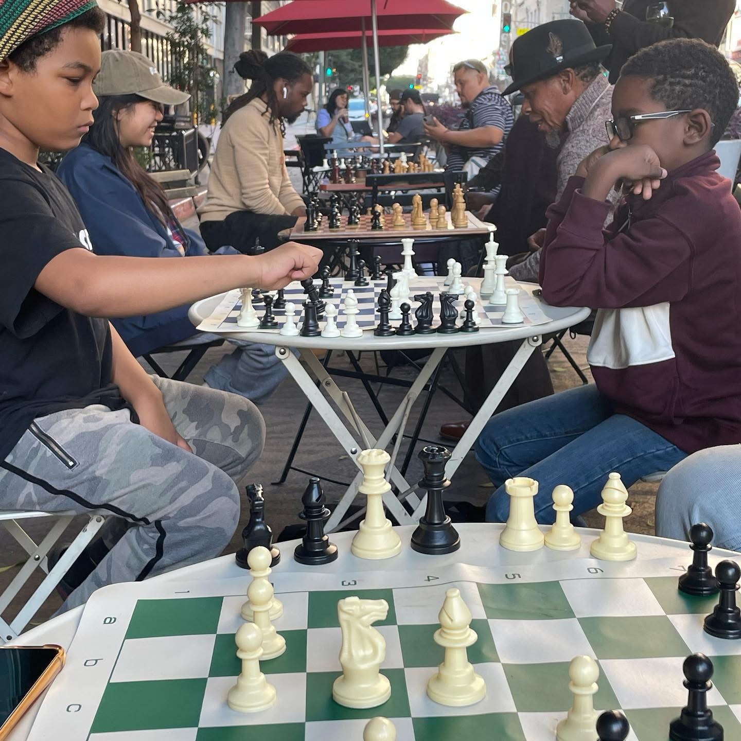 People playing chess outdoors at tables, with onlookers and city background, some wearing hats and sunglasses.