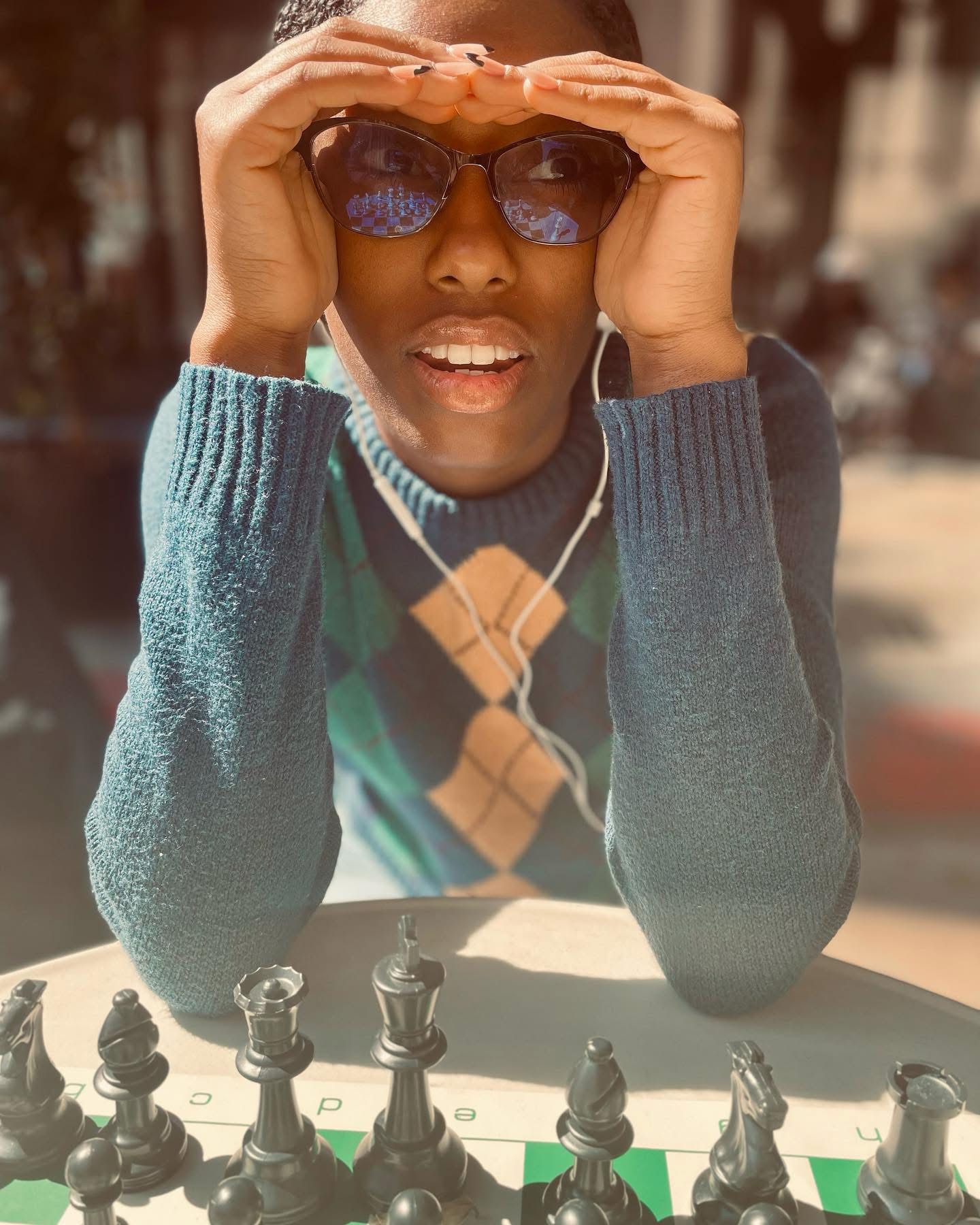 Person wearing sunglasses and a sweater, sitting at a table with chess pieces, outdoors on a sunny day.