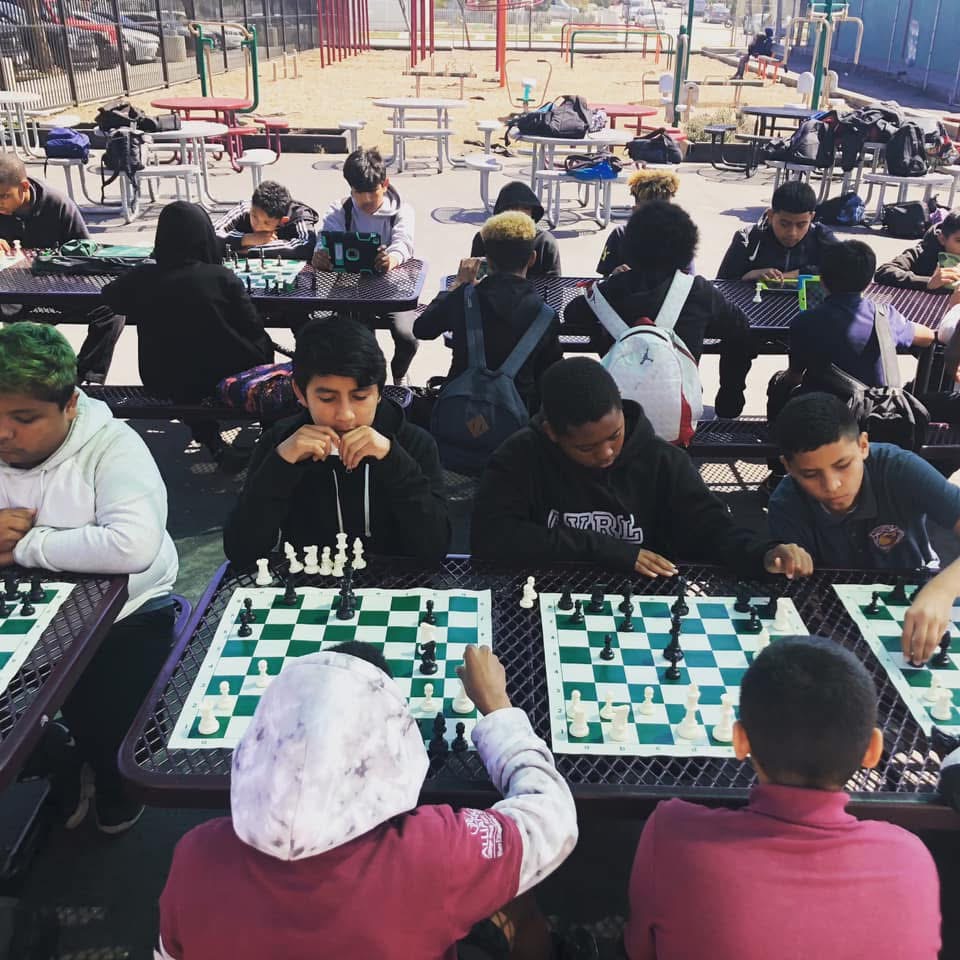 Group of children playing chess outdoors at tables, with playground equipment in the background.