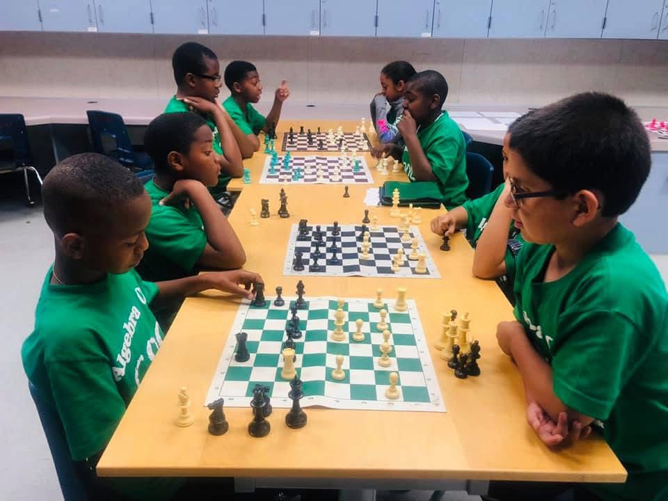 Children playing chess at a yellow table, wearing green shirts, in a room with a gray wall.