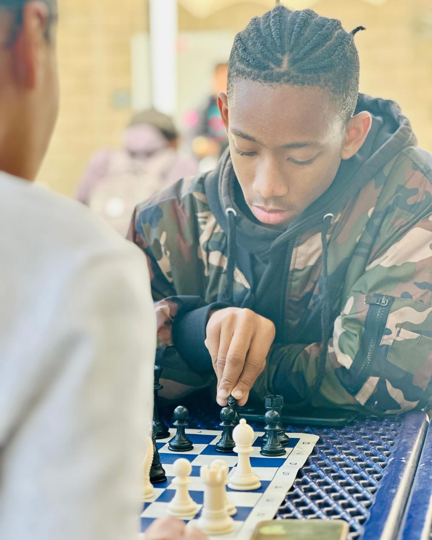 Young person with braided hair playing chess outdoors, focused on the game, with a blurred background.