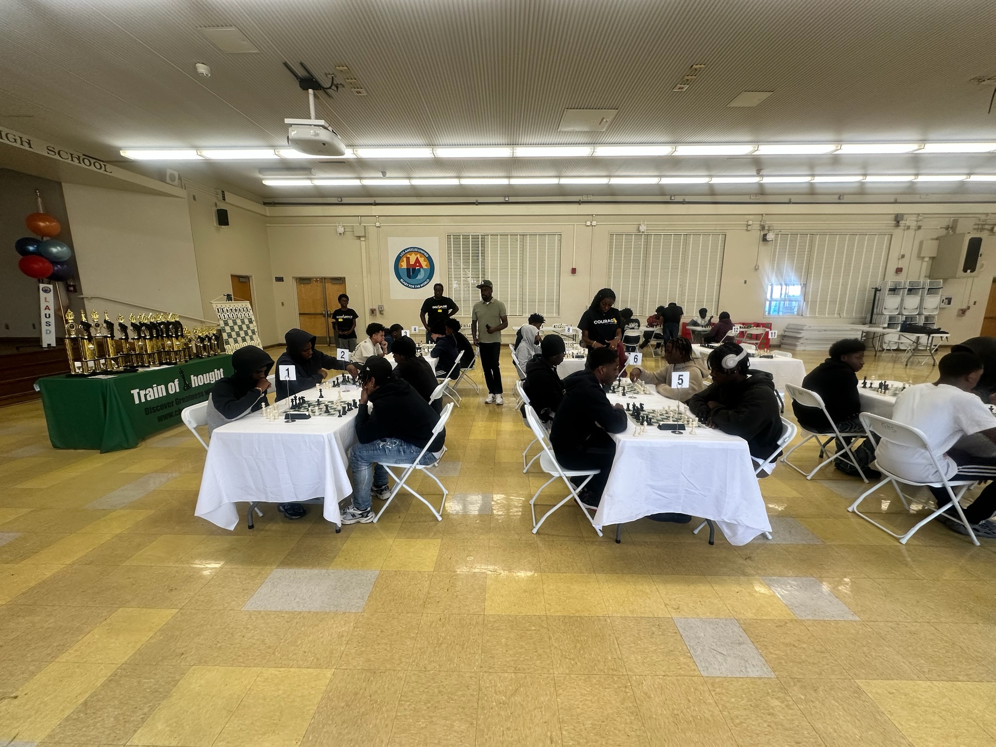 People seated at tables in a large room, possibly playing chess, with trophies on a table to the left.