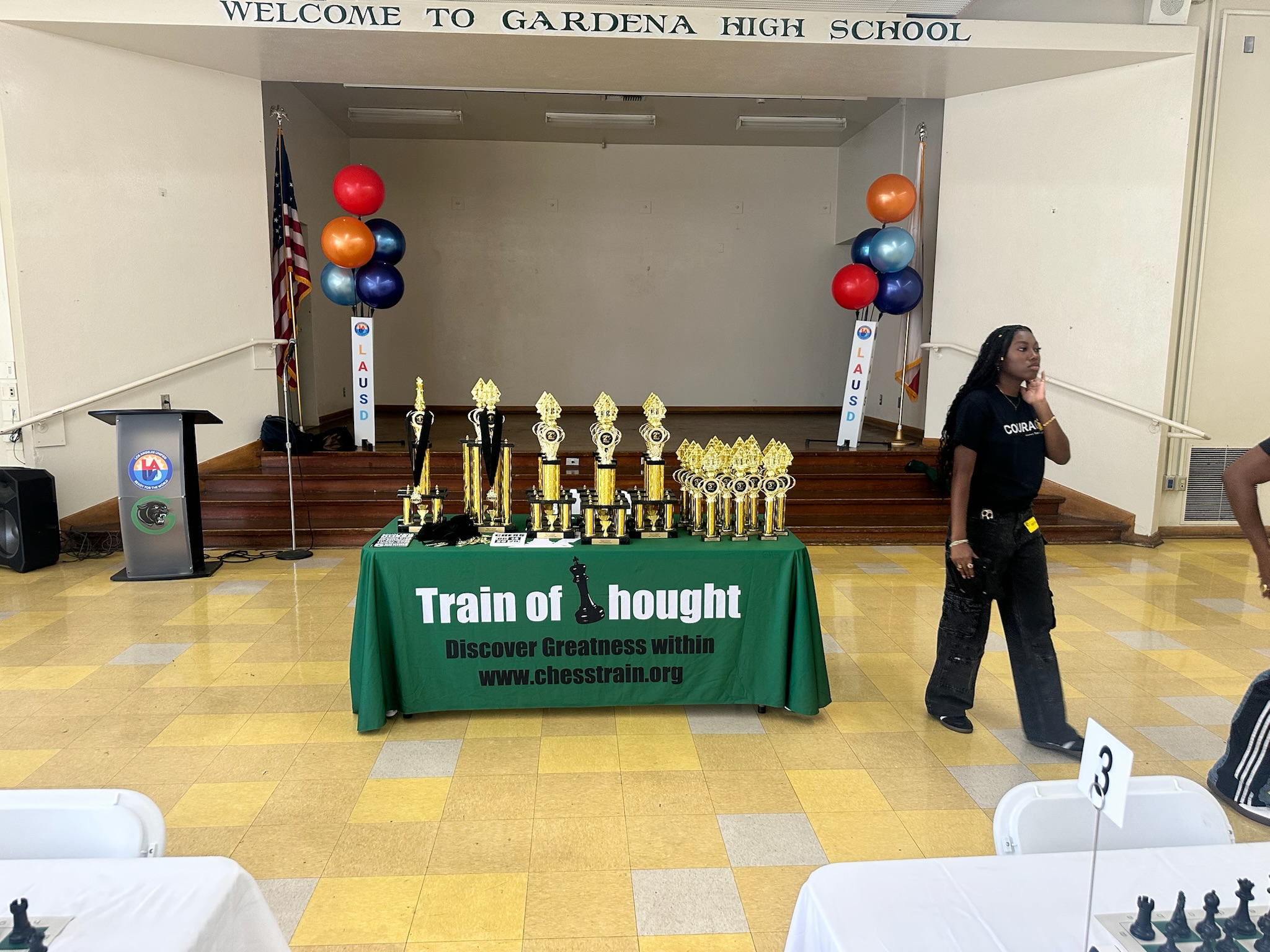 Awards table with trophies and balloons in a school gymnasium, with a woman walking nearby and a stage in the background.