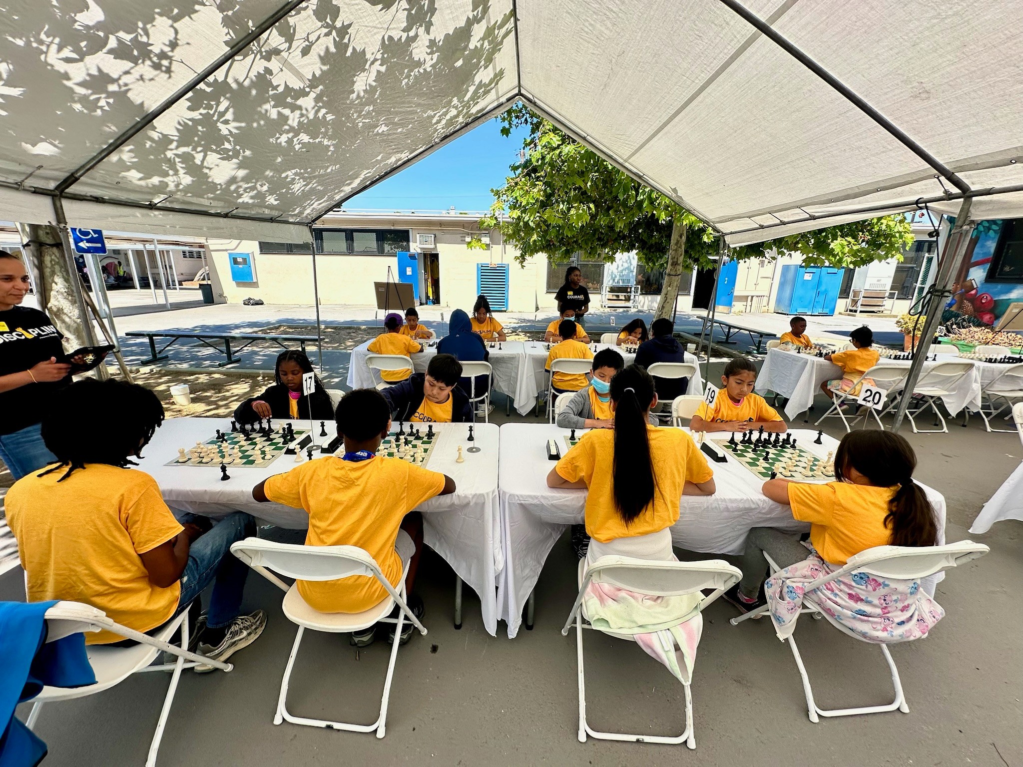 Children in yellow shirts playing chess under a large tent, with tables and chairs outside, trees and buildings in background.