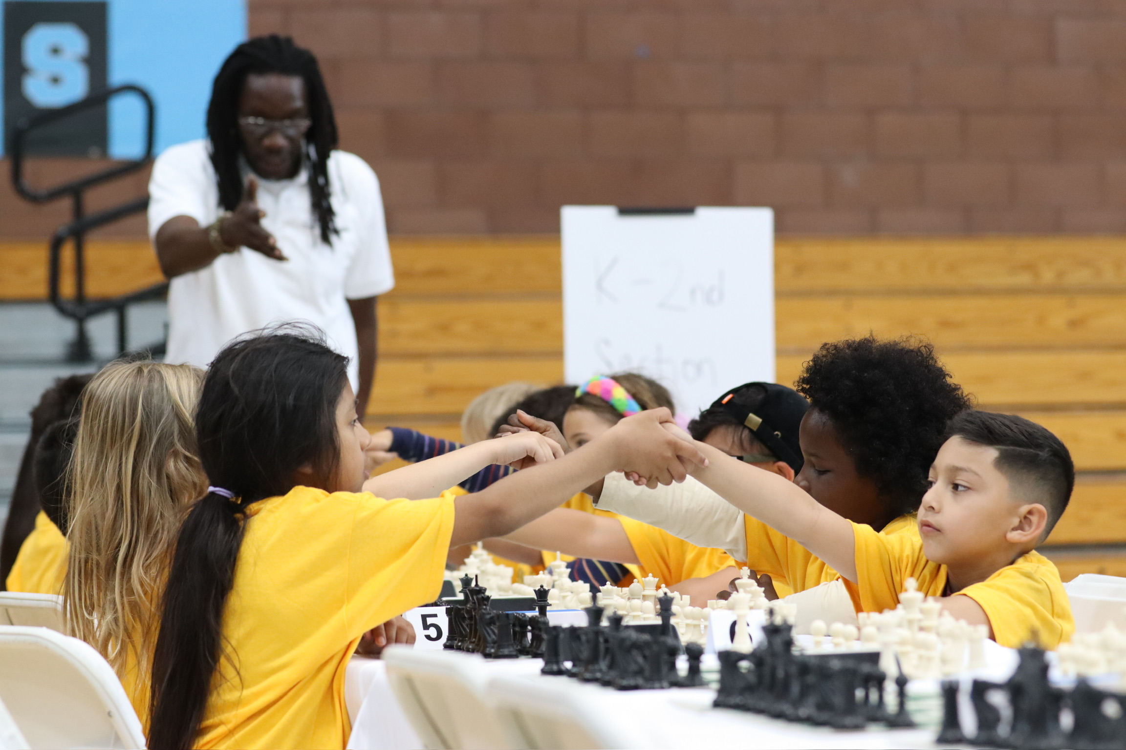 Children playing chess at a table, some wearing yellow shirts, in a school gymnasium with a brick wall background.