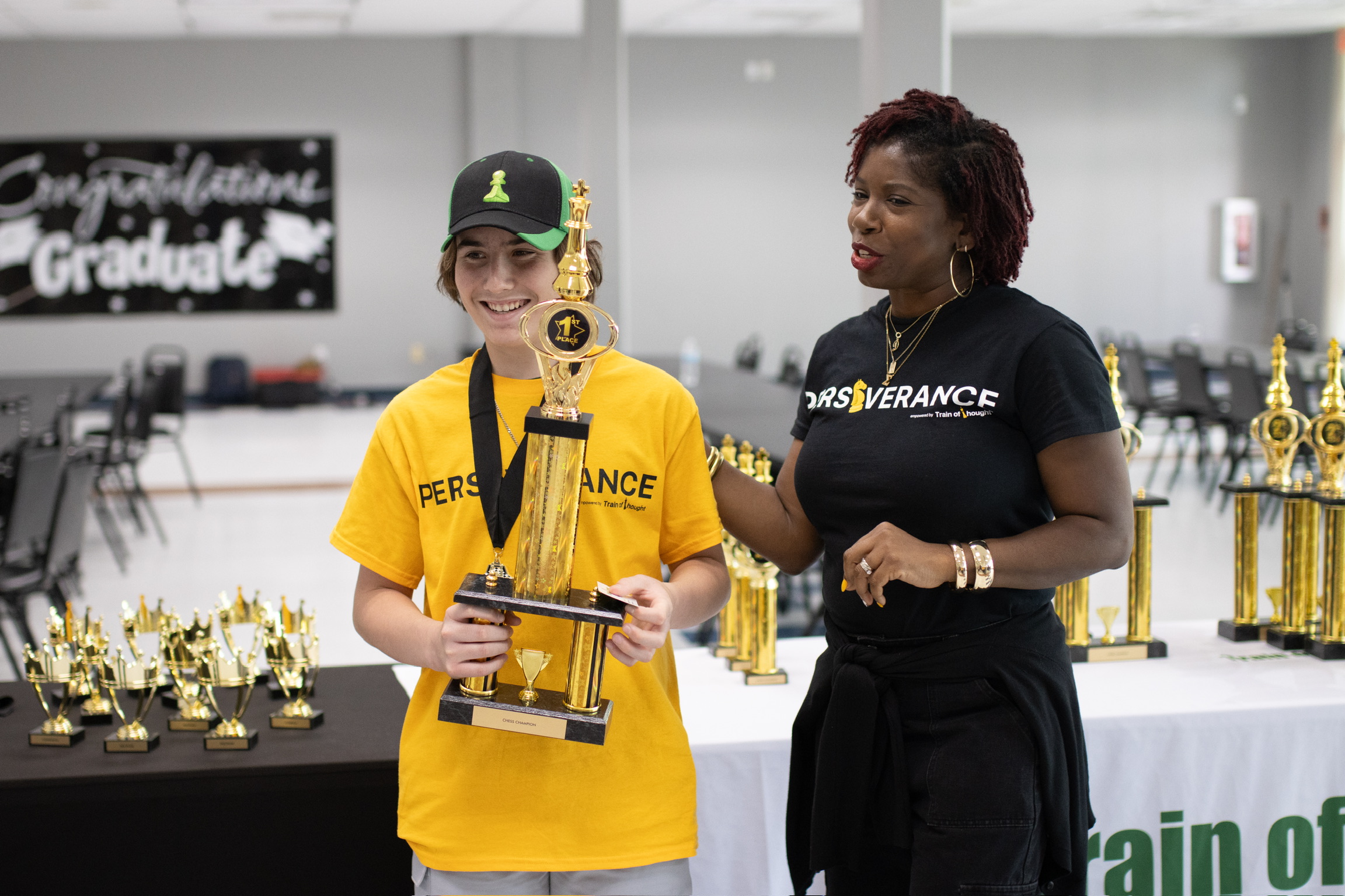 Two women standing indoors, one holding a trophy, with trophies on a table behind them, and a graduation sign in the background.
