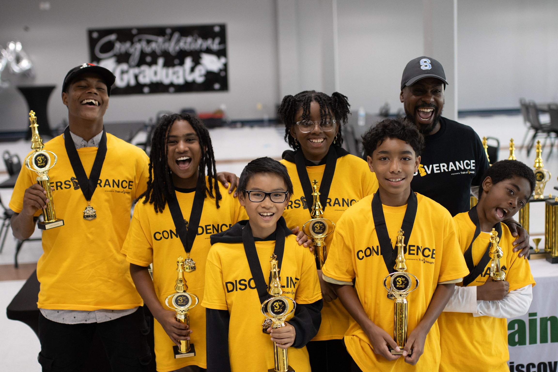 Group of six people in yellow shirts holding trophies, celebrating graduation indoors, smiling and posing for the photo.
