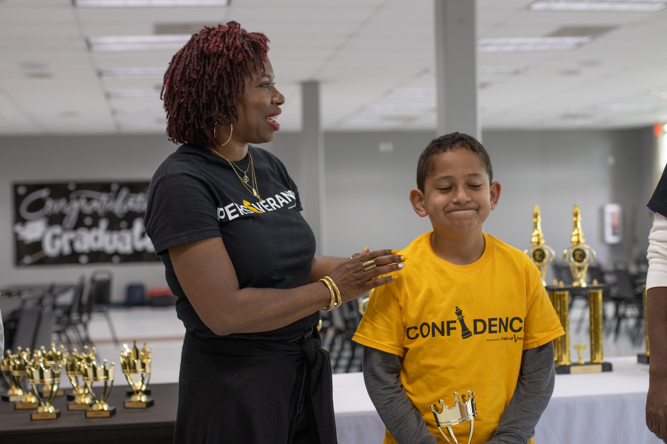 Woman and boy smiling, woman placing hand on boy's shoulder, trophies in background, both wearing yellow shirts, indoor setting.