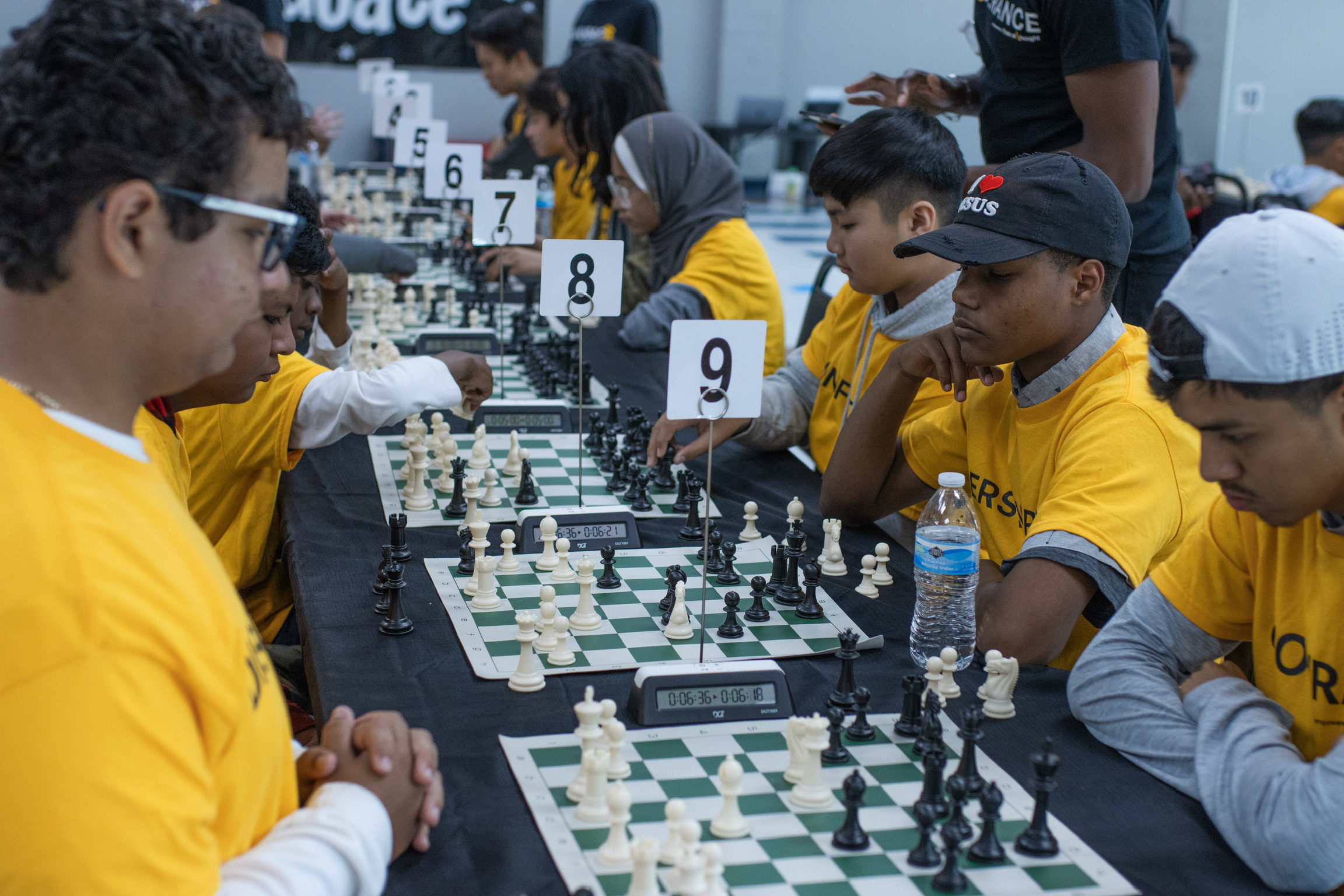 Children in yellow shirts playing chess at a tournament, seated at tables with numbered signs, water bottles nearby.