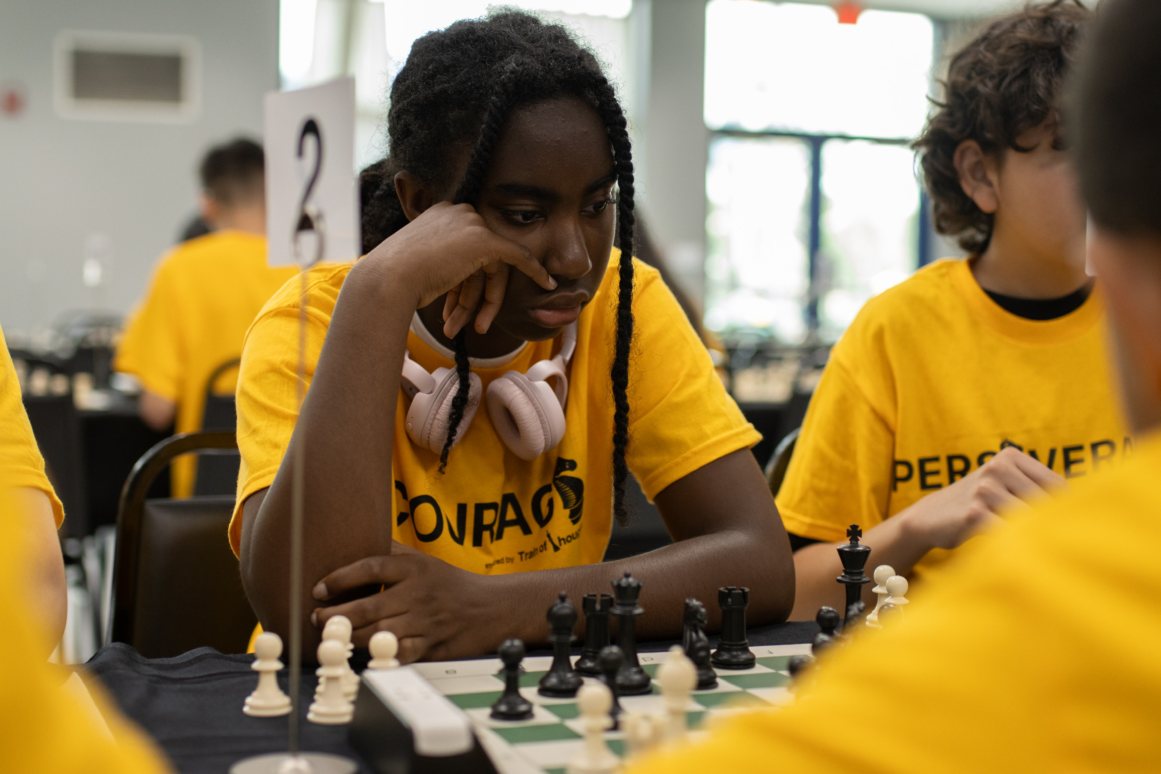 Girl with braided hair and headphones playing chess with others in a classroom, all wearing yellow shirts.