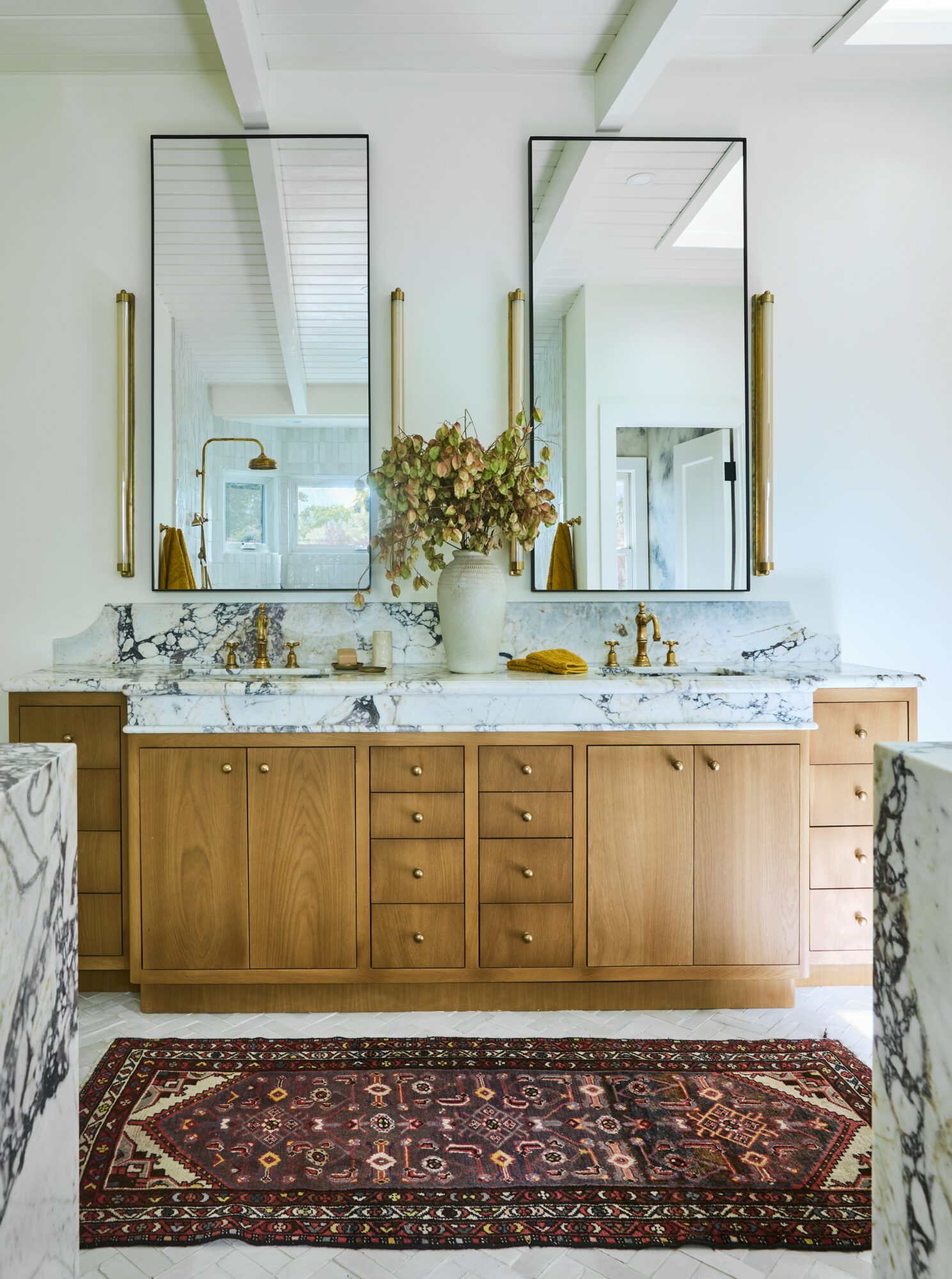 Bathroom vanity with two mirrors, marble countertop, wooden cabinet, and a rug in front.