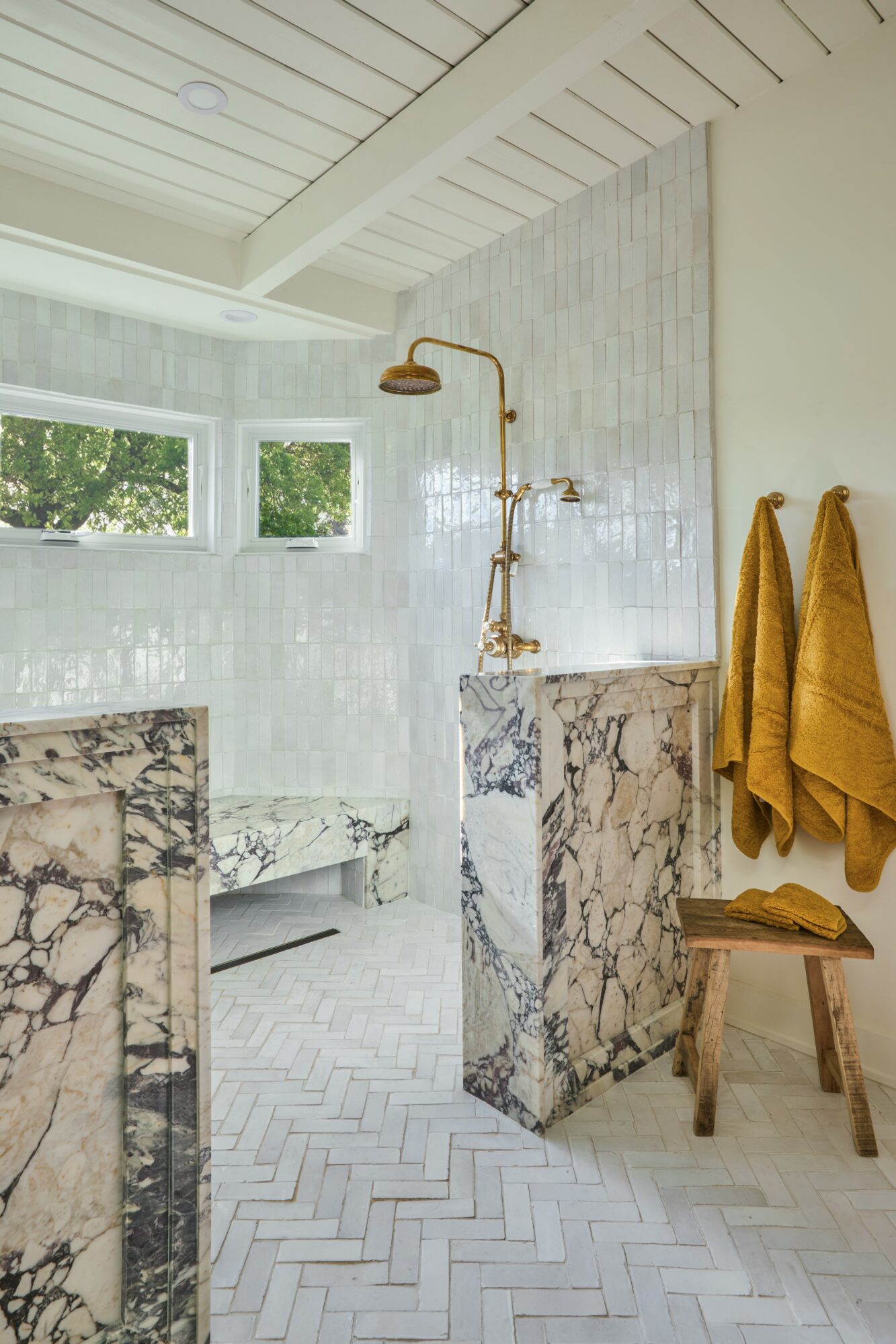 Bathroom with marble fixtures, a showerhead, and yellow towels hanging on a wooden stool, with windows and white tiled walls.