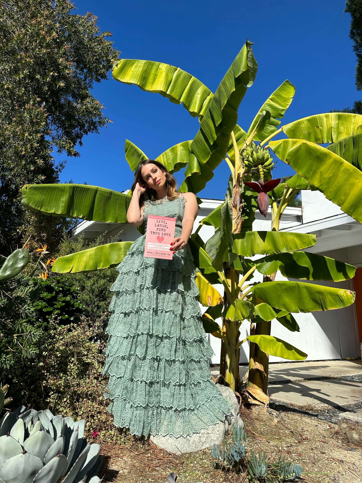Young woman in a long, layered dress stands outdoors among large banana leaves, holding a pink sign, with a clear blue sky.