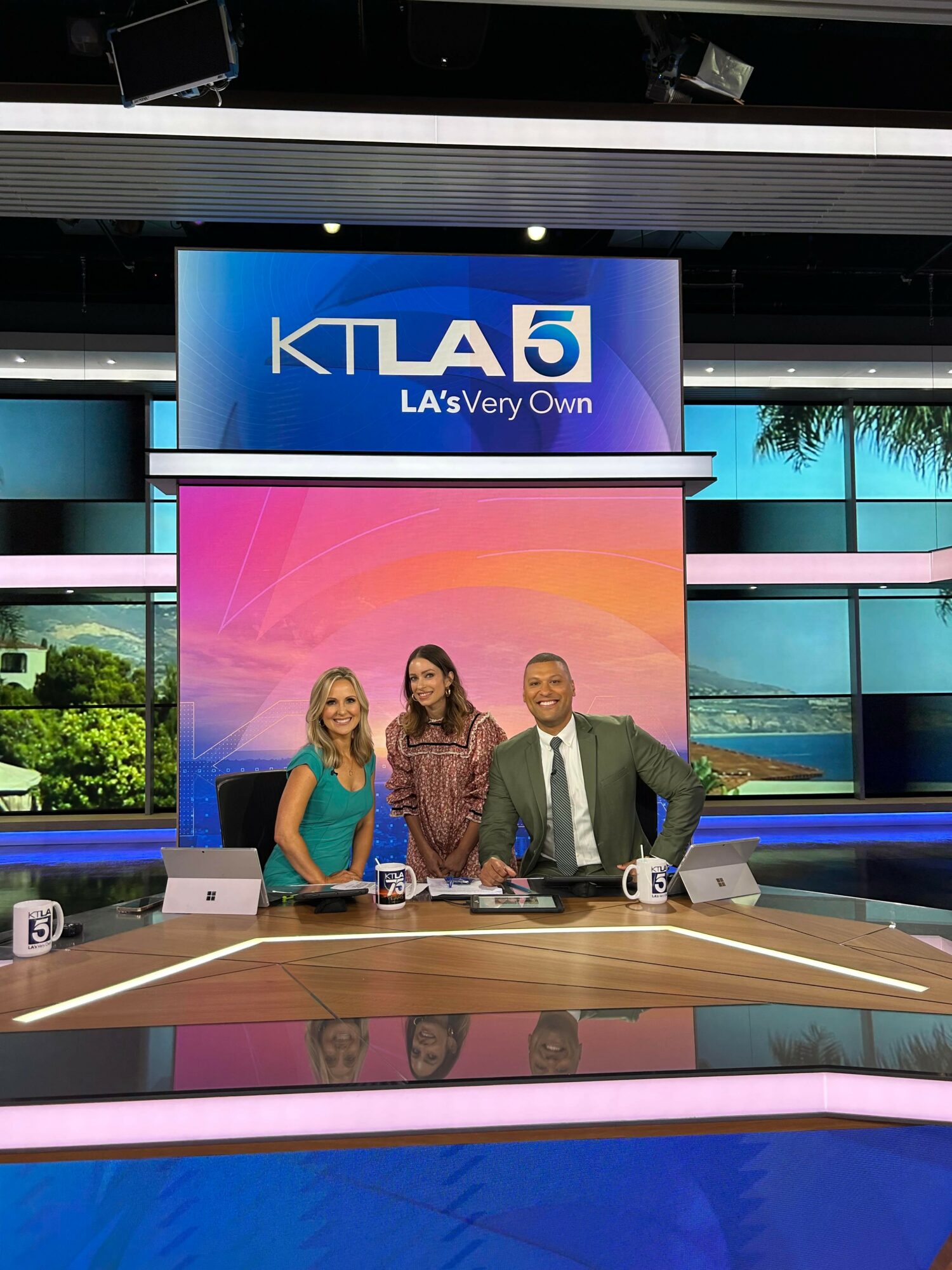 Three news anchors sit at a desk in a television studio with a large screen behind them displaying the KTLA 5 logo and a pink background.
