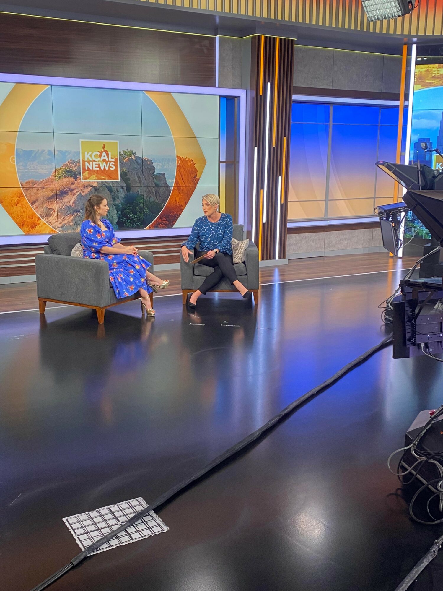 Two women sit on a gray couch and talk in a television studio with a large screen behind them displaying a news logo.