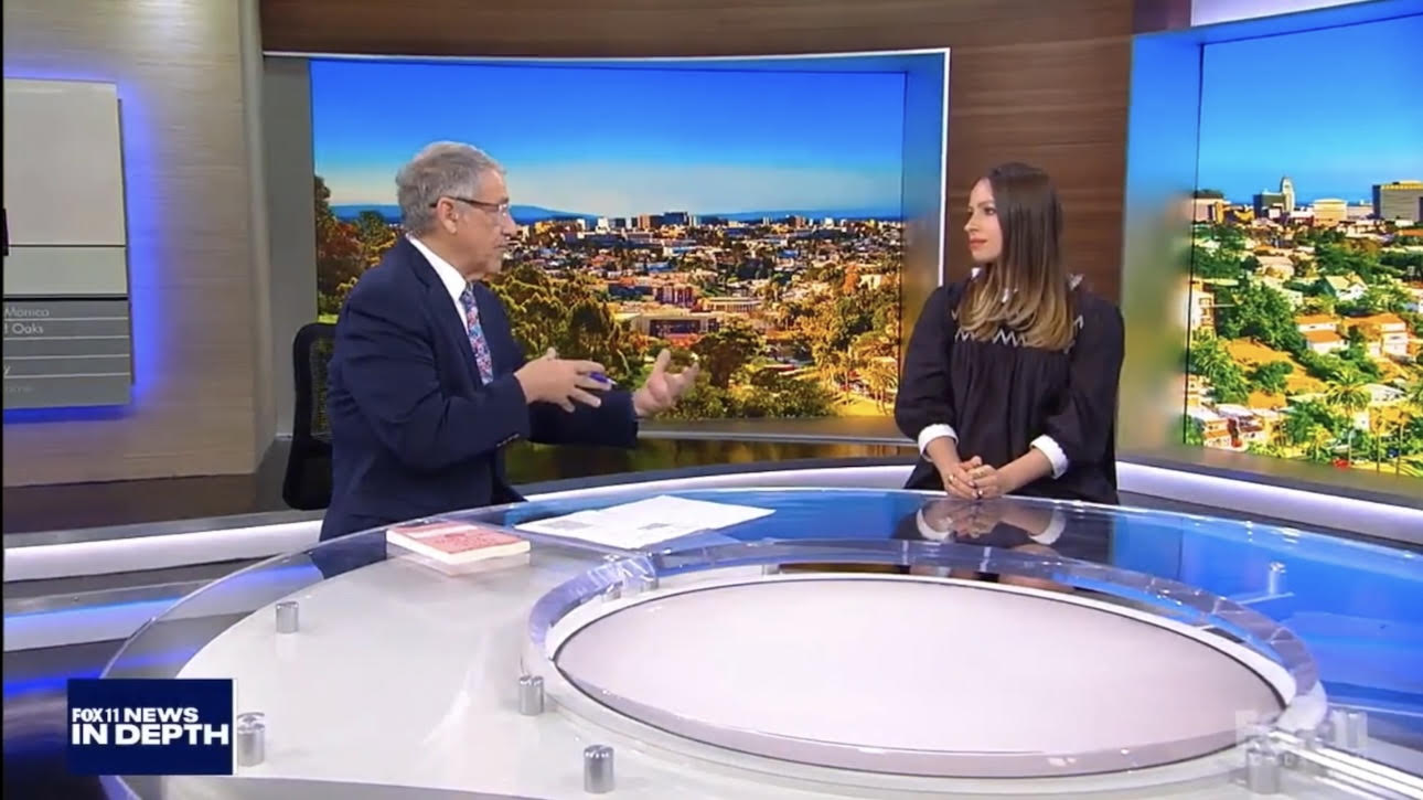 Two people sit at a news desk with cityscape screens behind them, engaged in conversation, in a television studio.