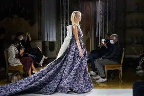 Woman in a long, flowing gown walking on a runway with seated audience and photographers, some wearing masks.