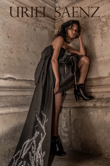 Young woman in black dress and high boots sitting on stone ledge, with a textured wall background.