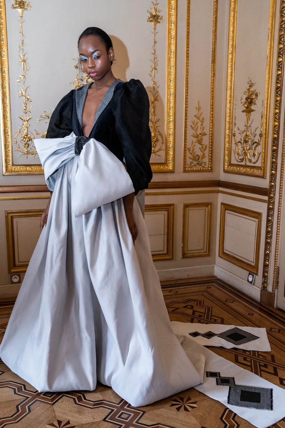 Woman in elegant gown with black top and large bow, standing in ornate gold-trimmed room, looking to the side.