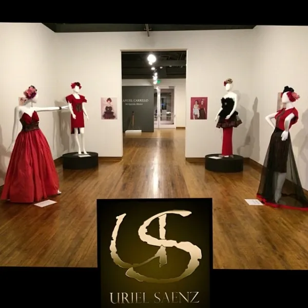 Exhibition room with mannequins dressed in red and black gowns, artwork on walls, wooden floor, and a black sign with a logo and text.