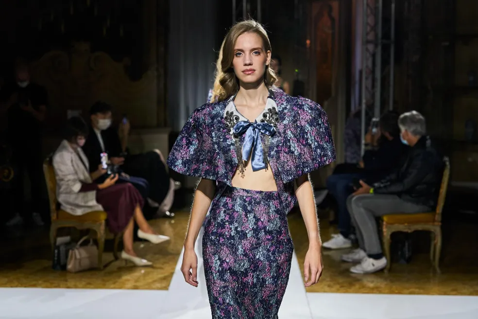 Fashion model walking on runway in floral outfit with puffed sleeves, audience seated in background, dim lighting.