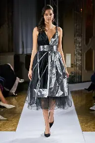 Woman walking on runway in black and silver dress with lace and metallic fabric, black heels, dark hair, in a fashion show setting.