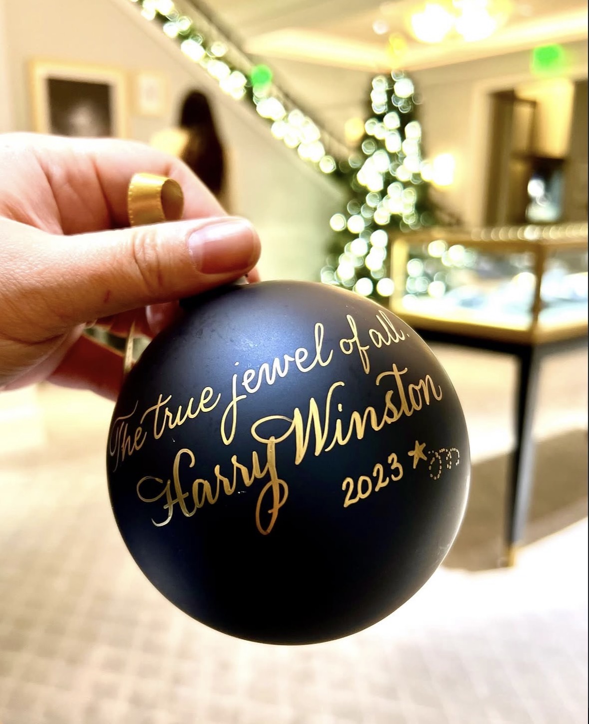 Hand holding a black ornament with gold writing, blurred indoor background with Christmas tree and lights.