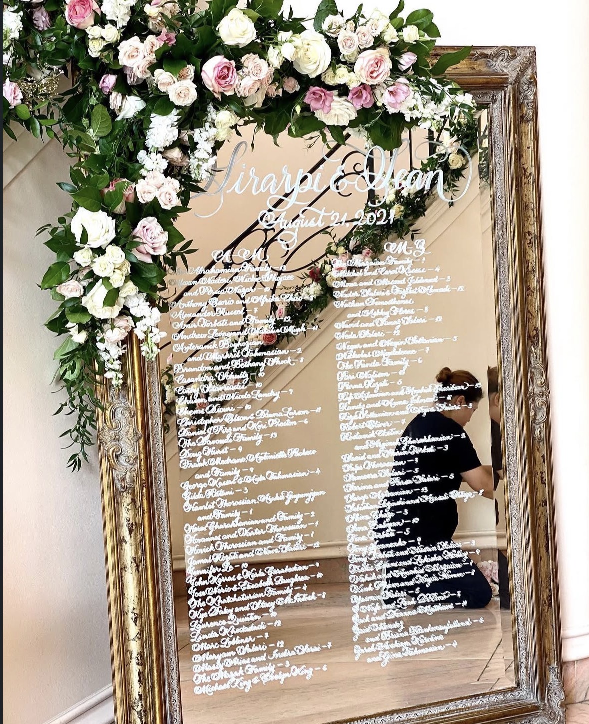 A mirror with floral decorations and a wedding schedule written on it, reflecting a person taking a photo.