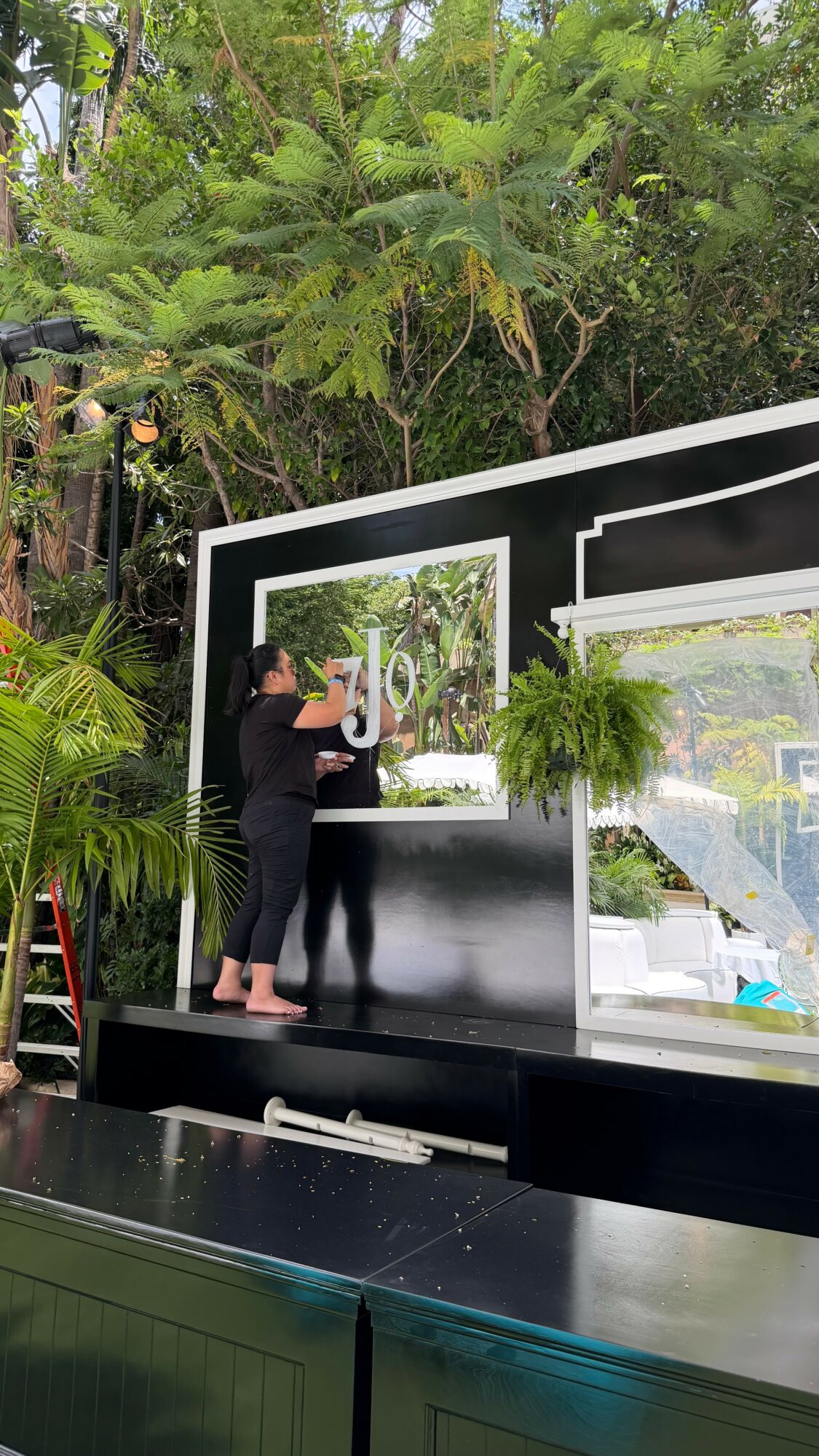 Person cleaning a large mirror on a black wall outdoors, surrounded by green plants and trees.