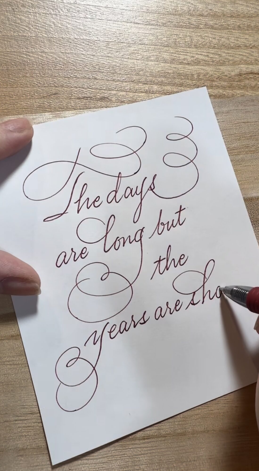 Hand holding paper with cursive writing on a wooden surface, with a pen in the bottom right corner.