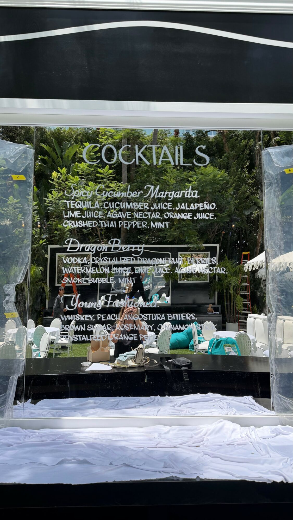 Menu board listing cocktails with ingredients and prices, reflected in a window with outdoor greenery in the background.
