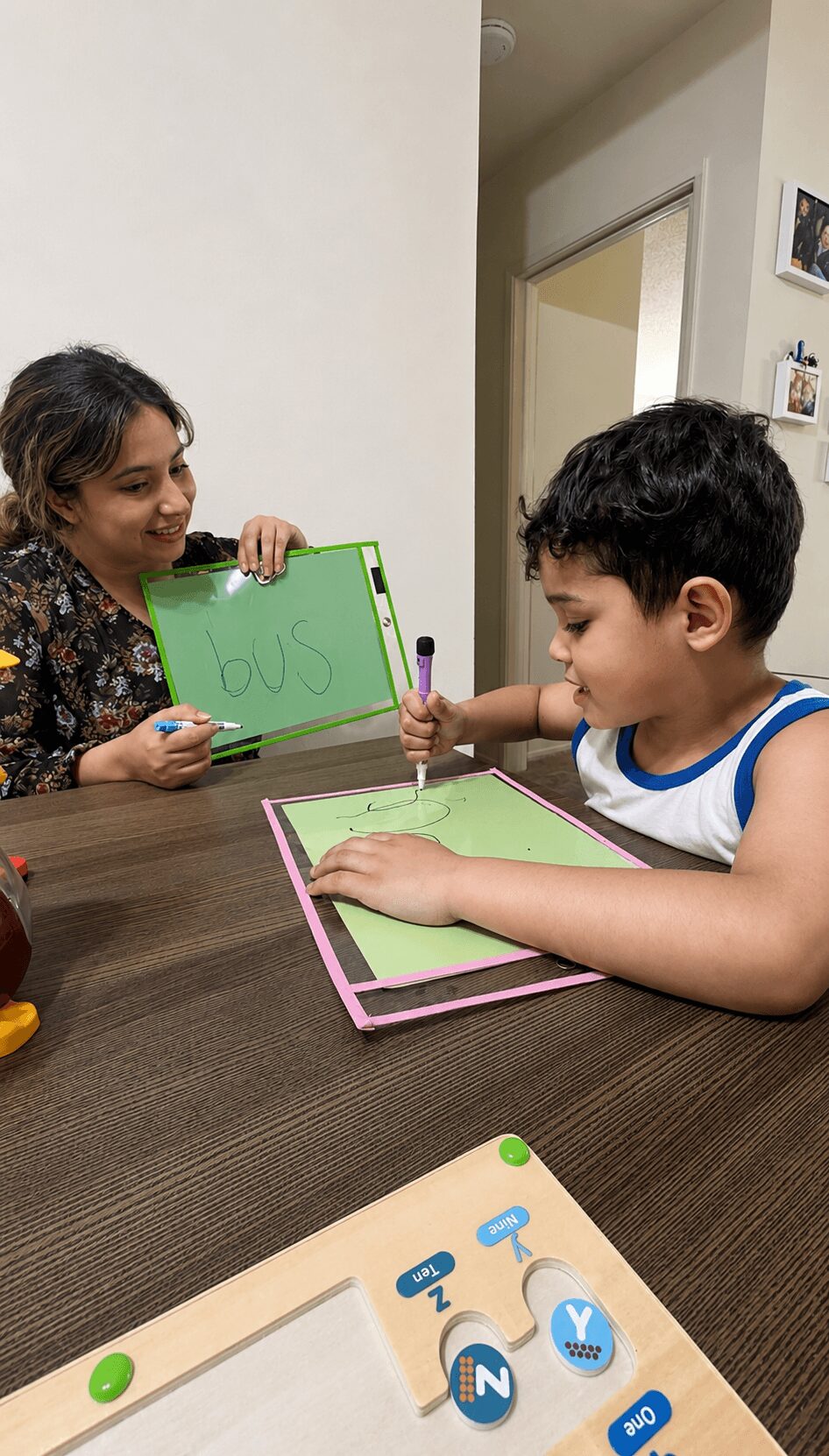 Child and woman sitting at a table, writing on small whiteboards, smiling, in a room with a door and pictures on the wall
