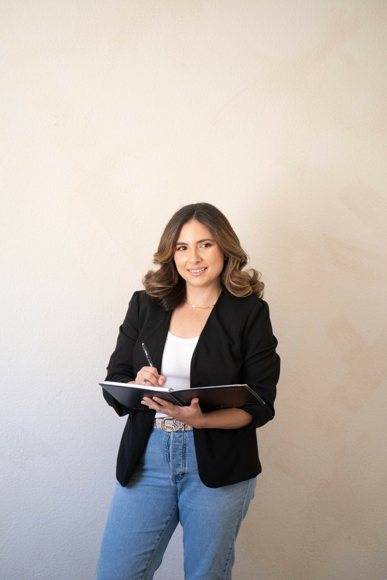 Woman with shoulder-length hair, wearing a black blazer and jeans, holding a notebook and pen, smiling.