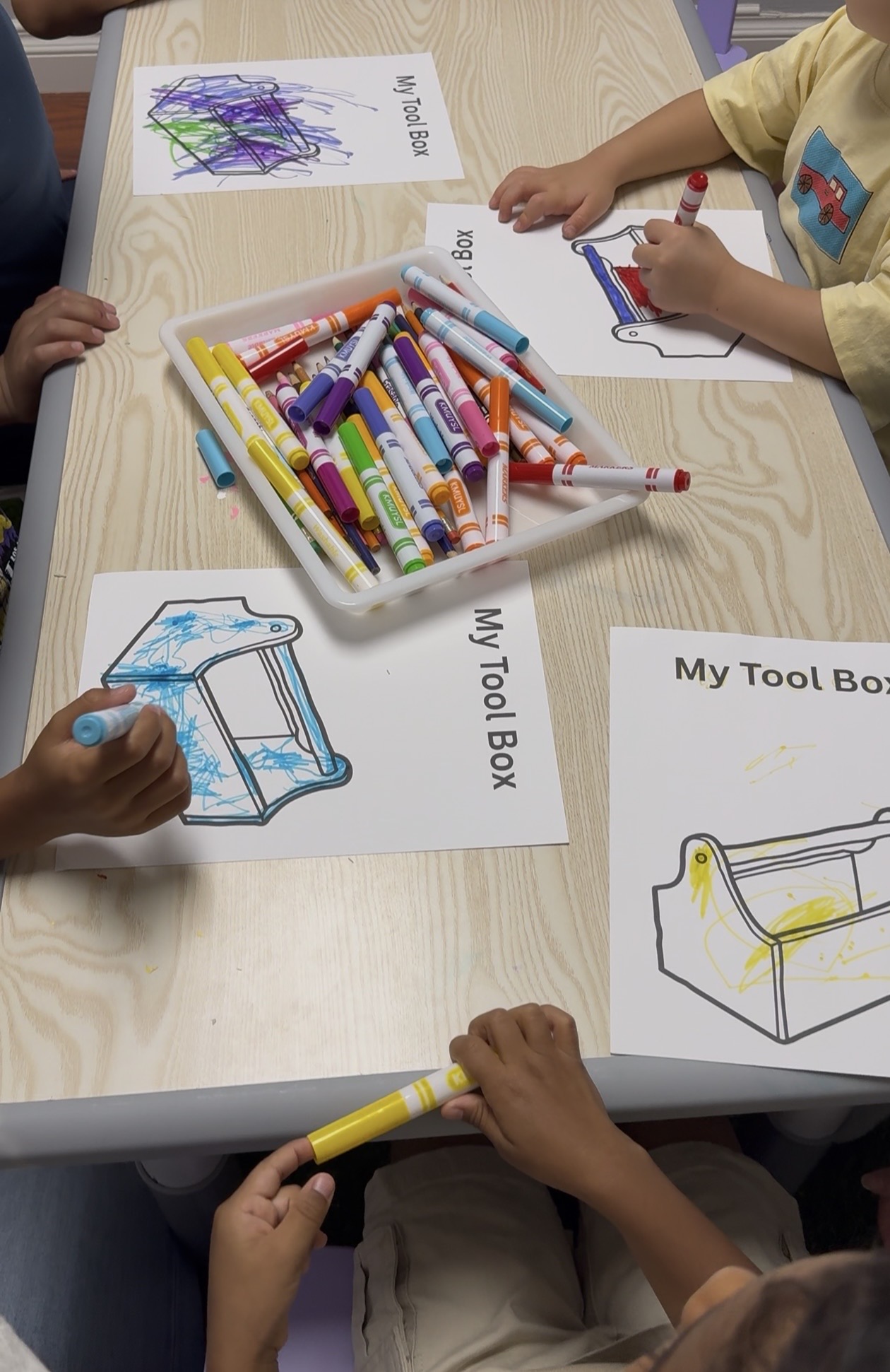 Children's hands drawing and coloring on paper with markers, with a box of markers and a drawing of a chair on the table.