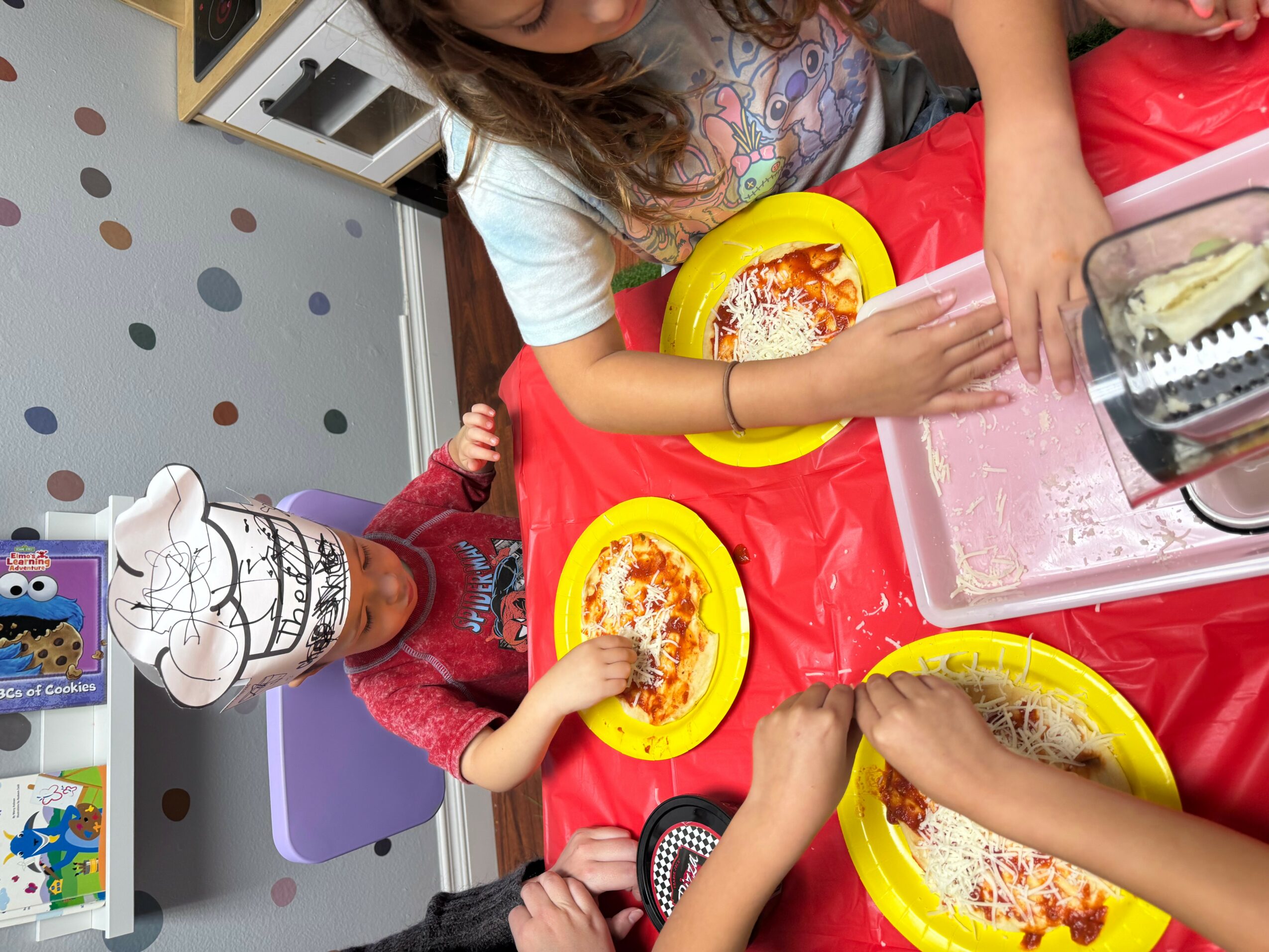 Children sitting at a table with pizza on yellow plates, one child wearing a paper chef hat, colorful background.