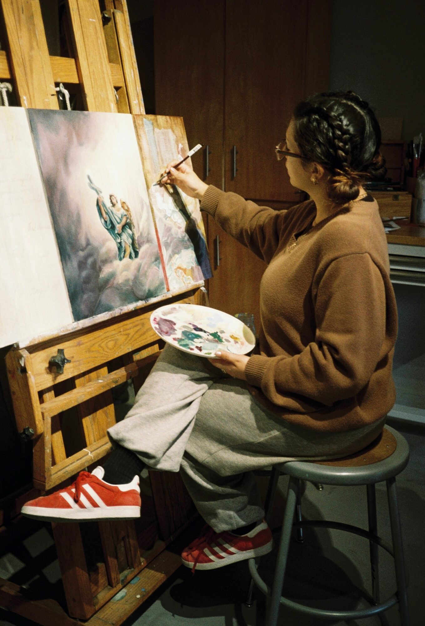 Person painting on canvas, seated on stool, holding a palette, wearing glasses, brown sweater, red and white sneakers.