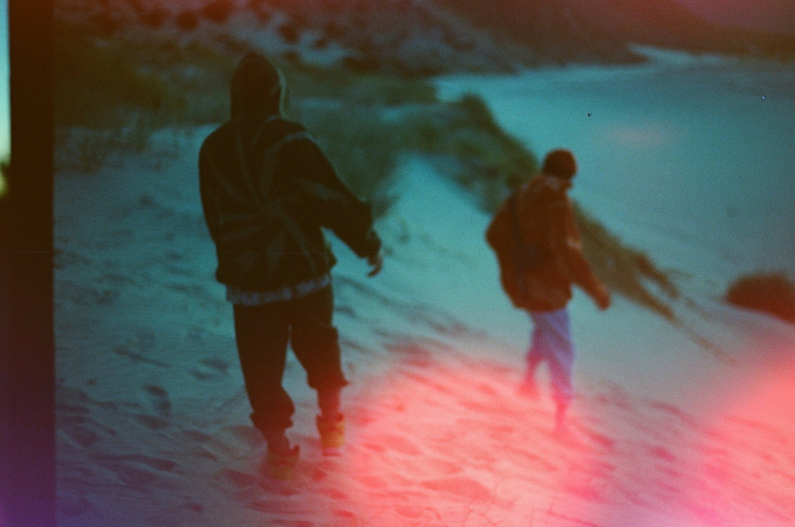 Two people walking on a snowy landscape near a body of water, with a blurred background and a pinkish light reflection.