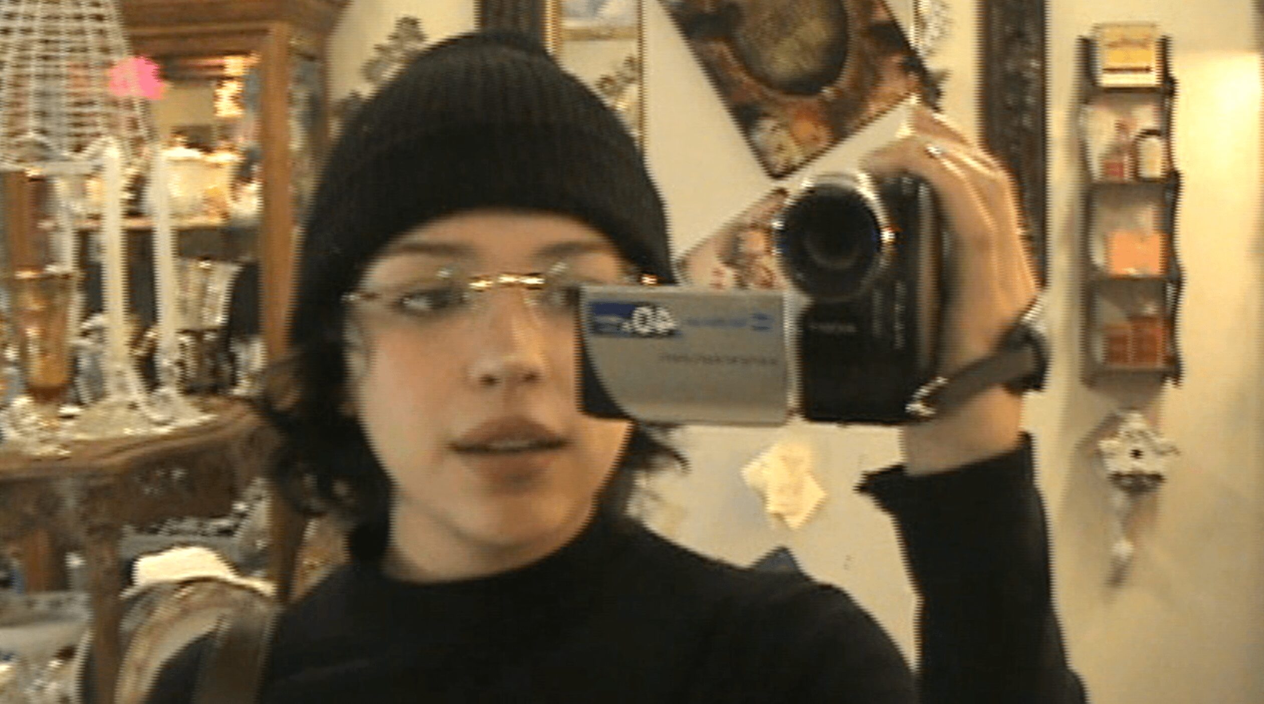 Person wearing glasses and a black beanie taking a mirror selfie in a room with shelves and decorations