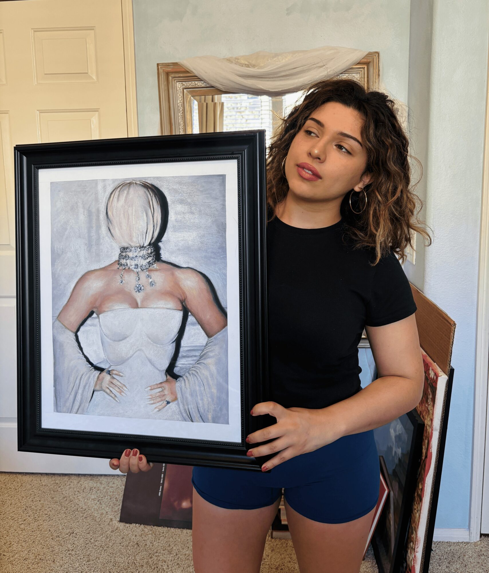 Young woman with curly hair holding a framed portrait of a woman with short hair and jewelry, standing in a room.