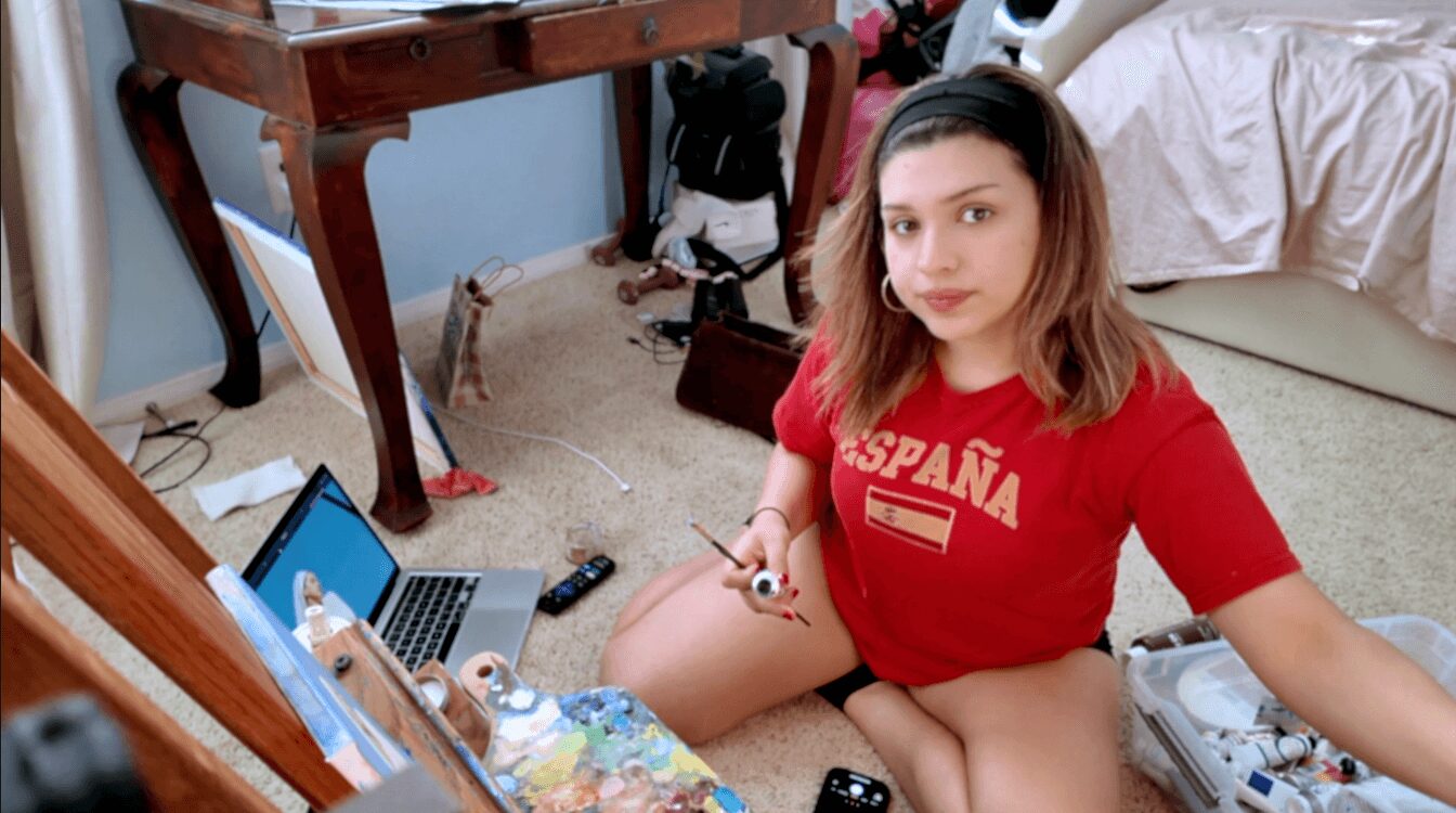 Young woman sitting on the floor in a cluttered room, wearing a red T-shirt, surrounded by various objects.